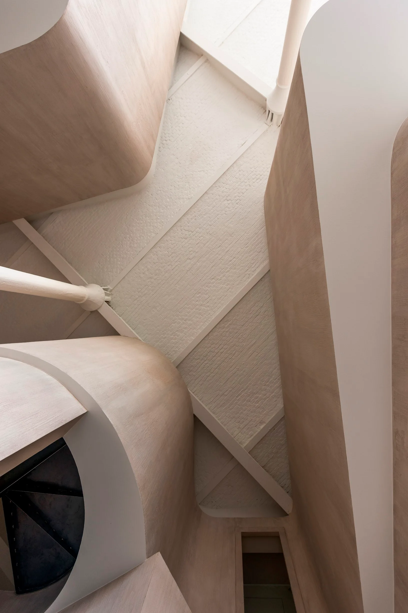 An upward view of a modern stairwell featuring curved beige walls, white structural columns, and exposed ceiling beams against a textured brick ceiling.