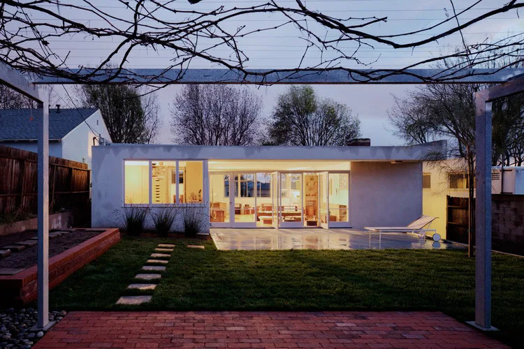 A mid-century modern home exterior at twilight, featuring a glowing glass wall, a concrete patio with a lounge chair, a green lawn, and foreground brick paving.