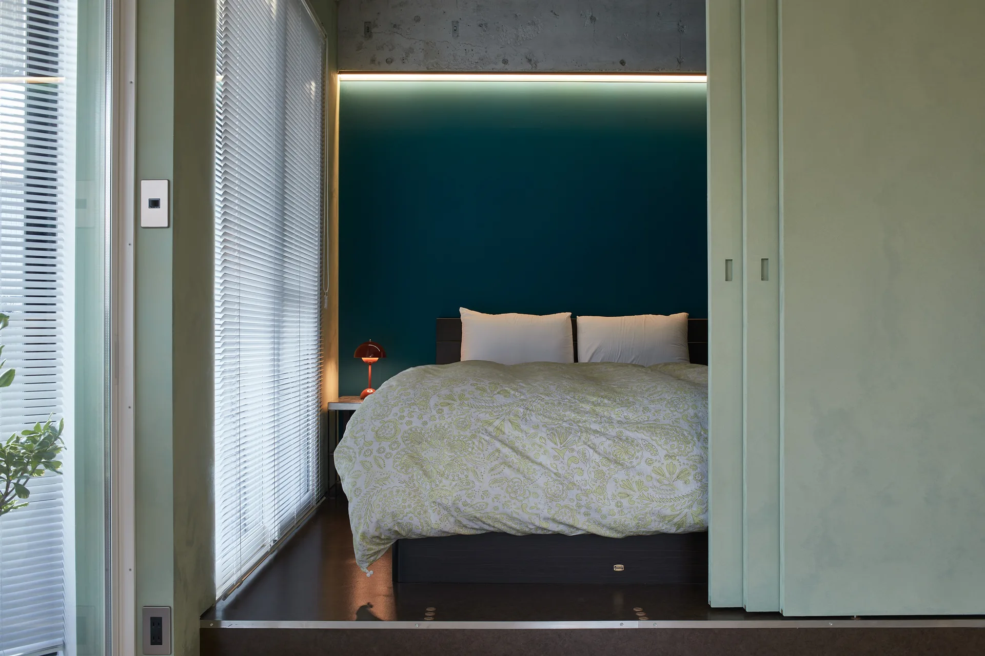 A raised platform bed with a floral duvet sits against a dark teal accent wall, illuminated by cove lighting below an exposed concrete ceiling.