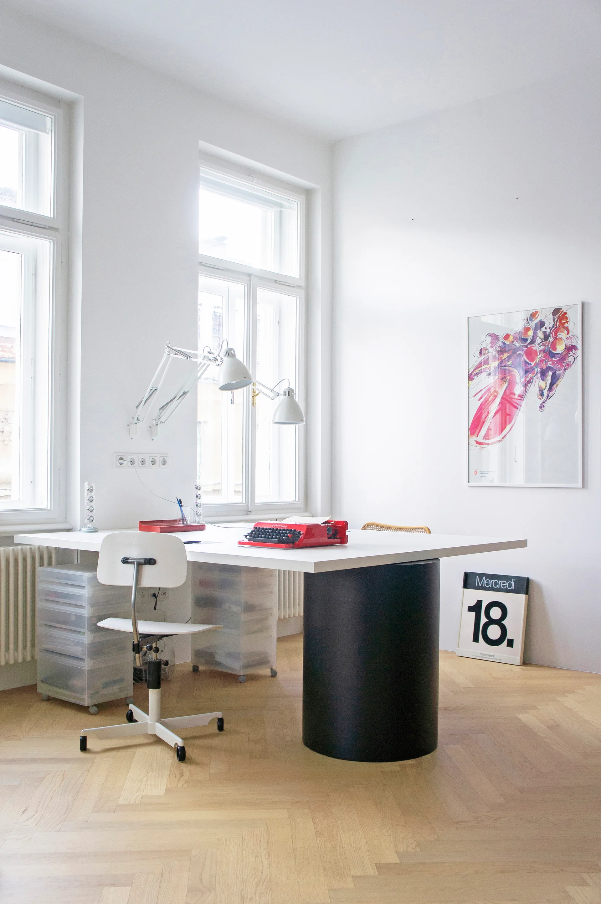 A minimalist home office features a white desk supported by a black cylinder leg, a white office chair, and herringbone hardwood floors.
