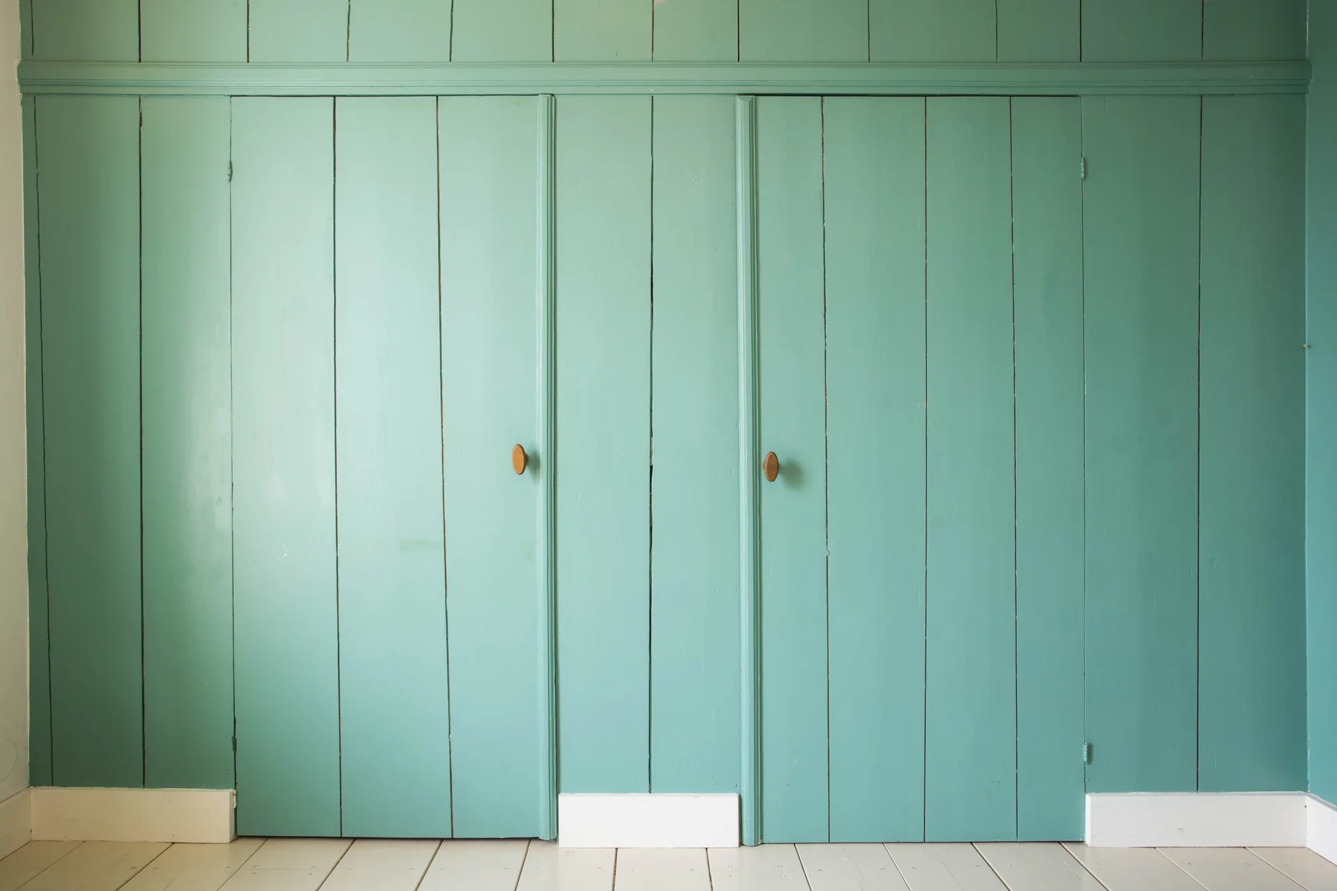 A wall of vertical wood planks painted in a muted teal shade features integrated closet doors with small wooden knobs and a white floor.