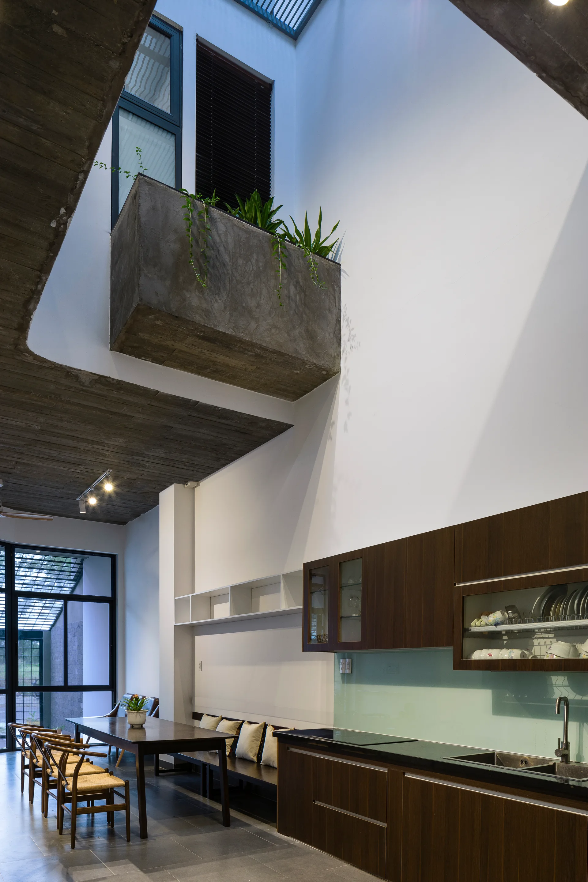 A modern kitchen and dining area with dark wood cabinetry, a dining bench, and an elevated concrete planter box under a high white ceiling.
