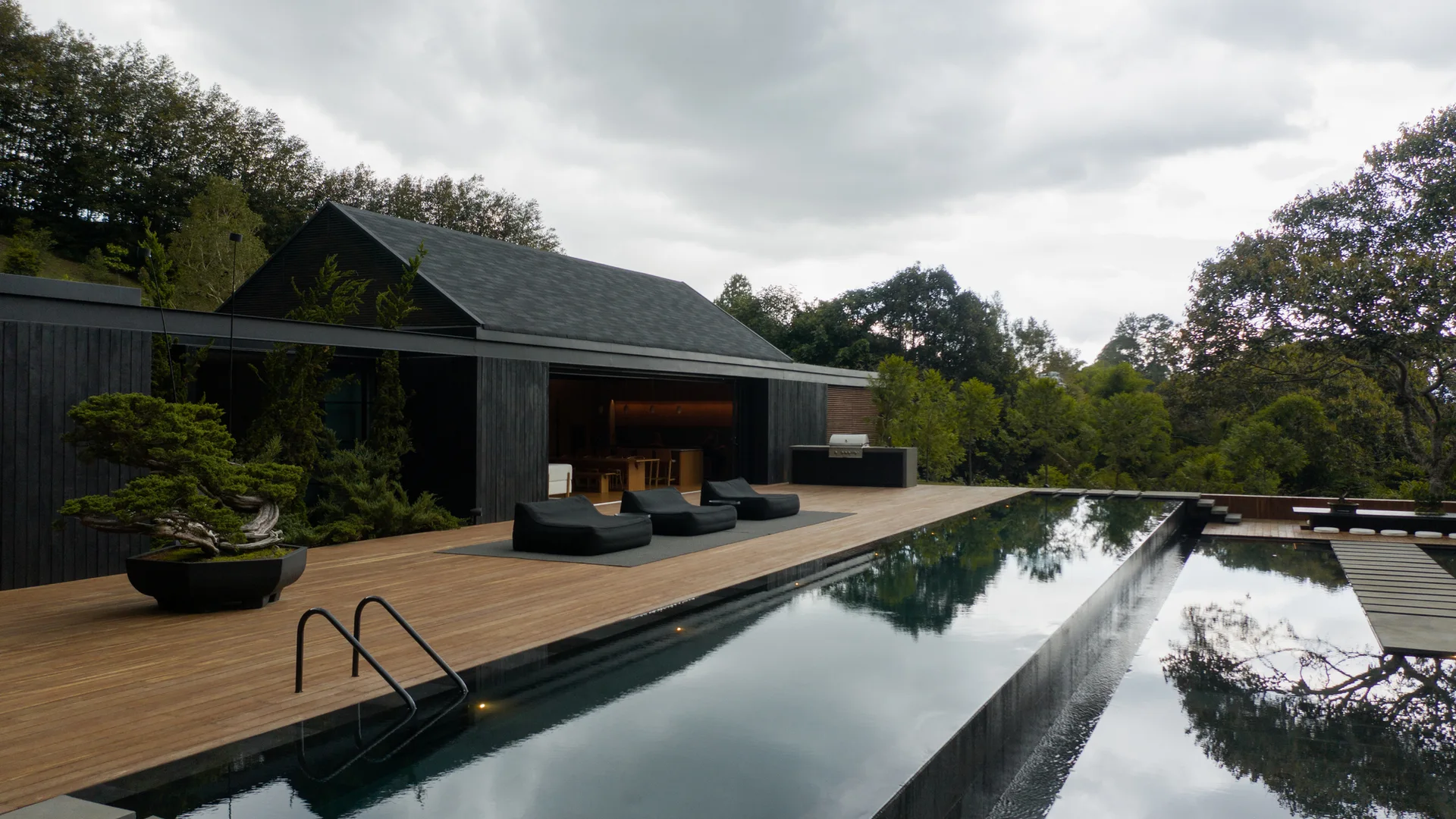 A modern charcoal-clad house features a wide wooden deck with black lounge chairs next to a large reflective infinity swimming pool.