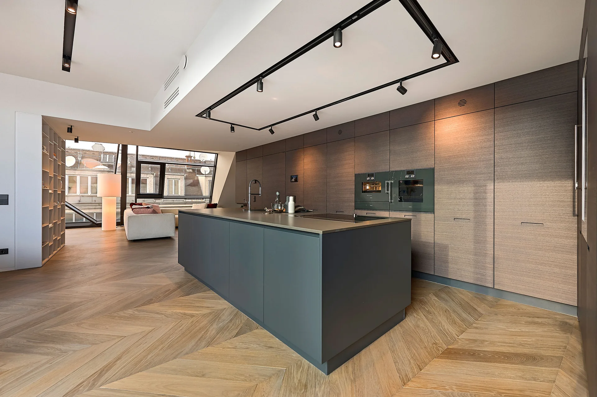 A modern kitchen featuring a dark grey island, floor-to-ceiling wood cabinetry, and herringbone wood floors beneath black track lighting.