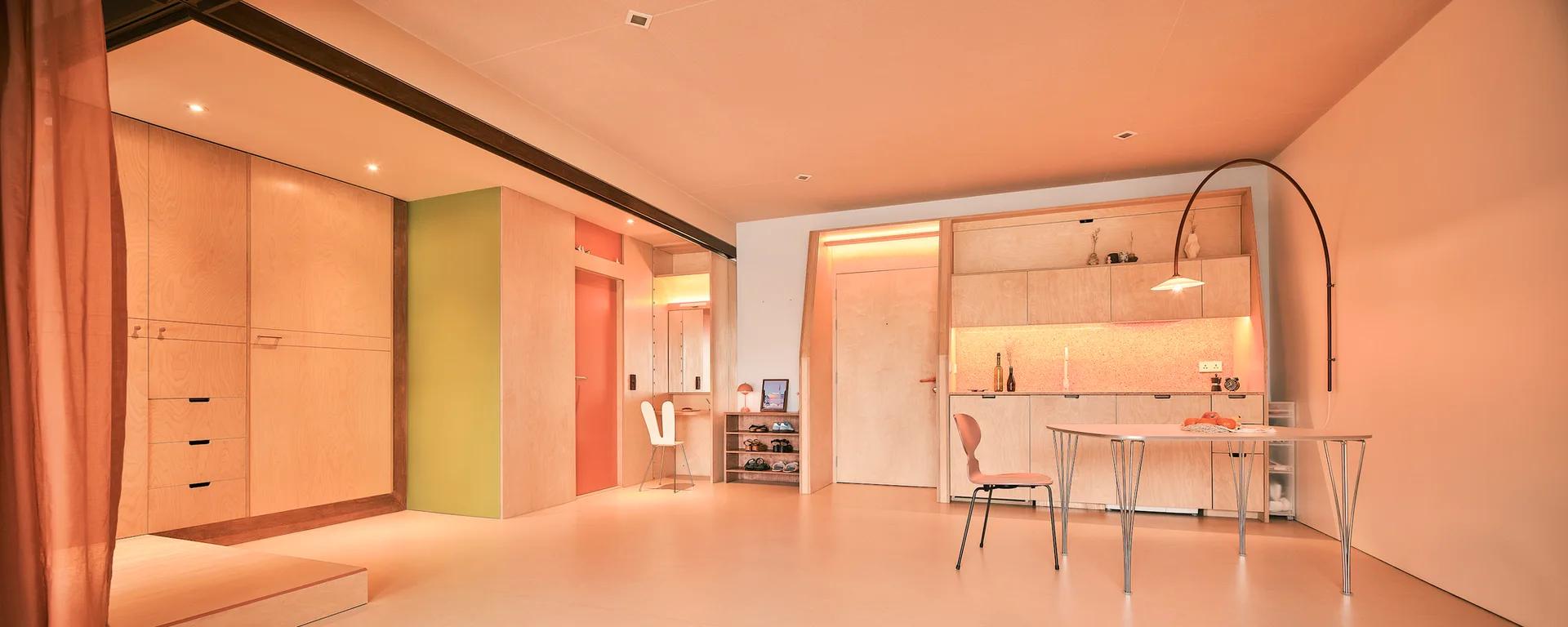 A minimalist open-plan kitchen and dining area featuring custom plywood wall cladding, matching wood cabinetry, and a curved floor lamp.
