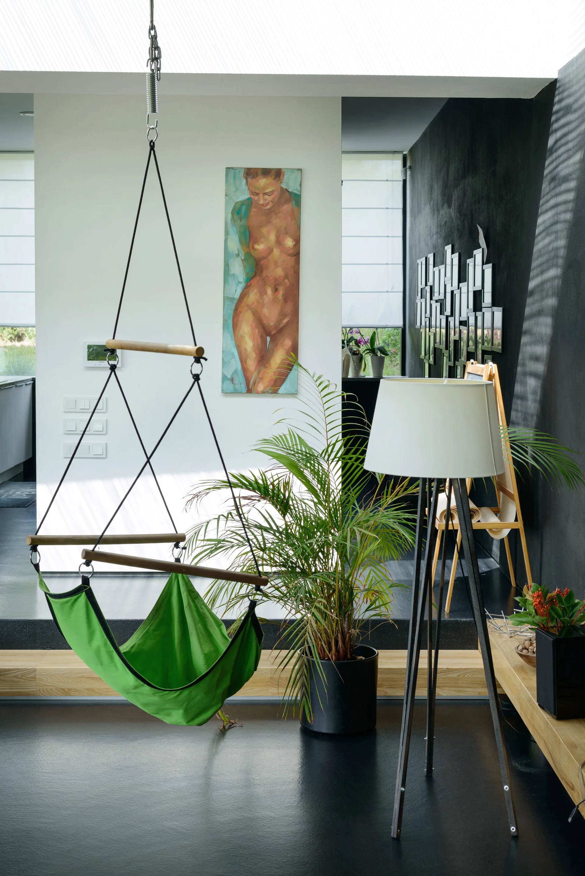 A modern interior features a bright green hanging hammock chair, a tall white tripod lamp, a potted palm, and contrasting black and white walls.