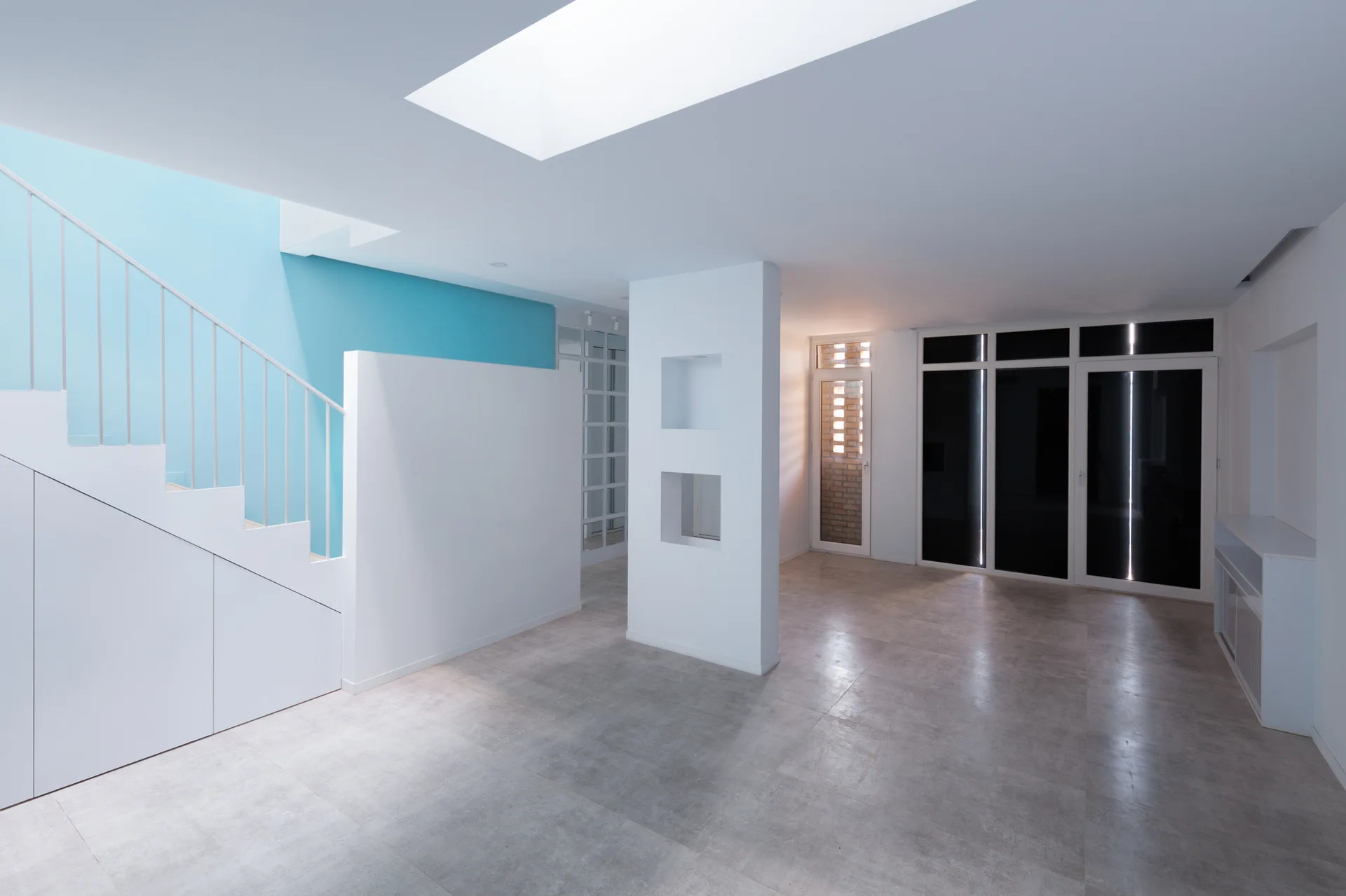 A minimalist white staircase features a bright cyan accent wall, grey tiled flooring, a skylight, and a central pillar with square niches.