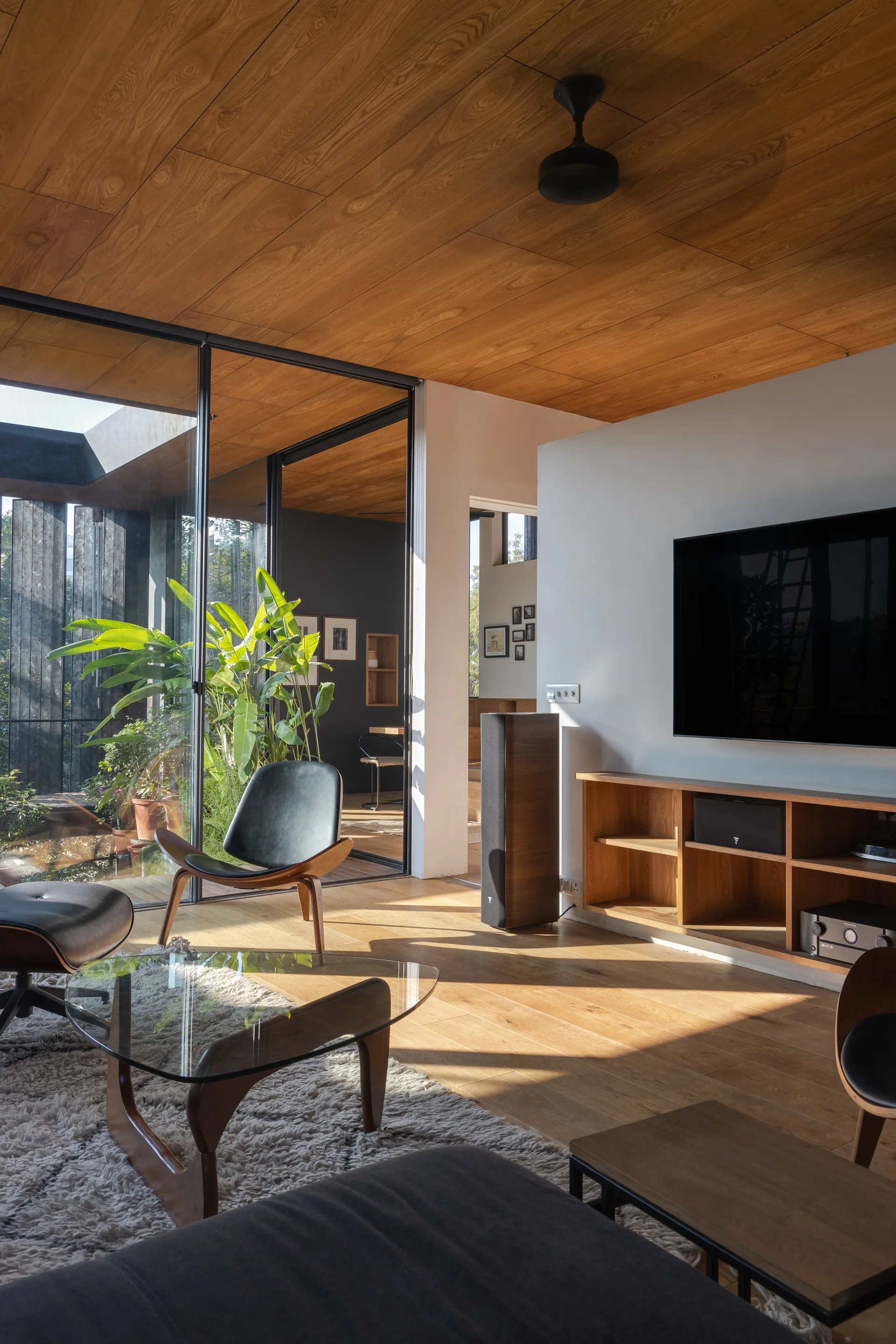 A mid-century modern living room featuring wood plank ceilings, leather lounge chairs, and sliding glass doors that open to a green courtyard.