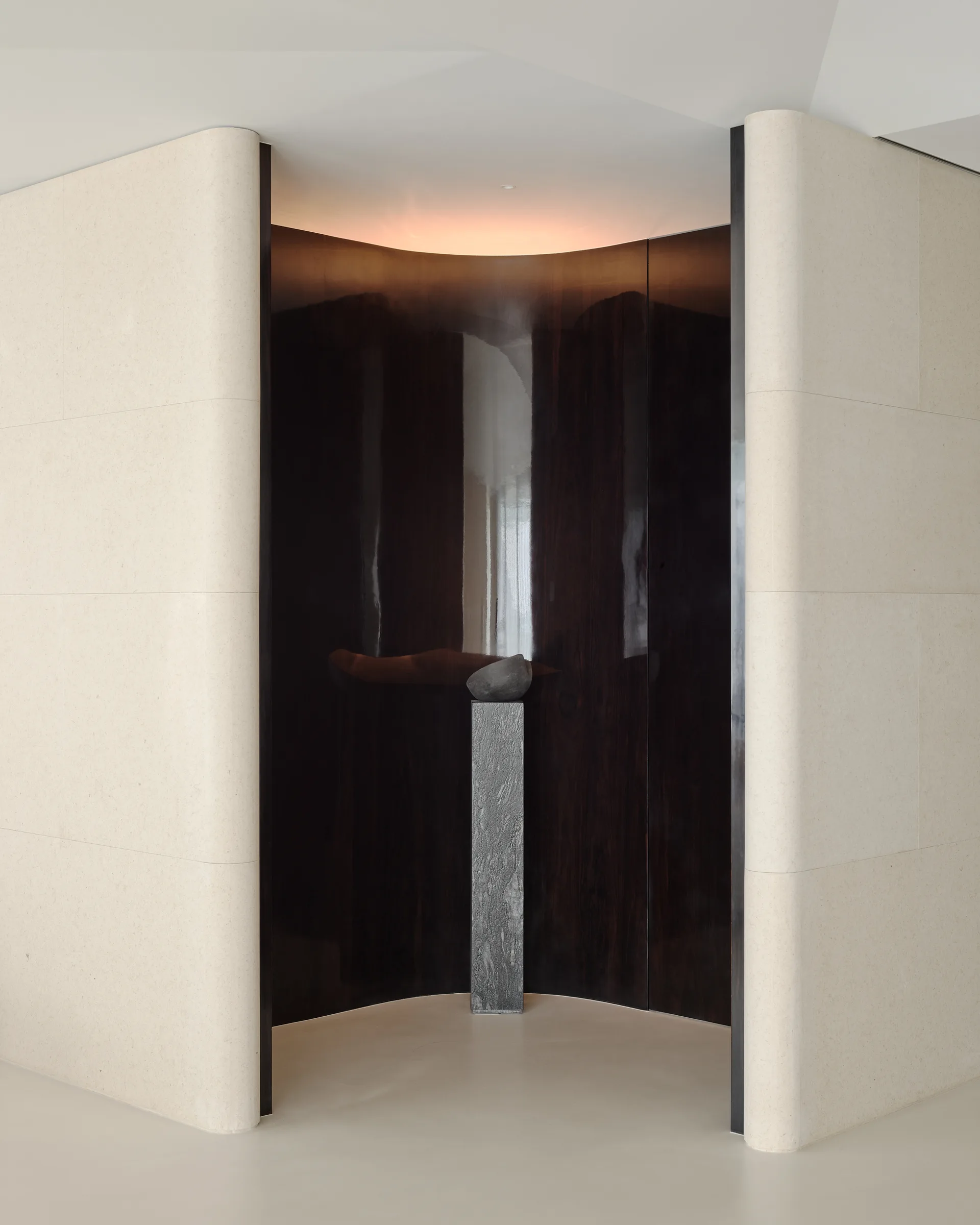 A textured stone pedestal with a small sculpture sits inside a curved dark wood niche flanked by beige stone walls.
