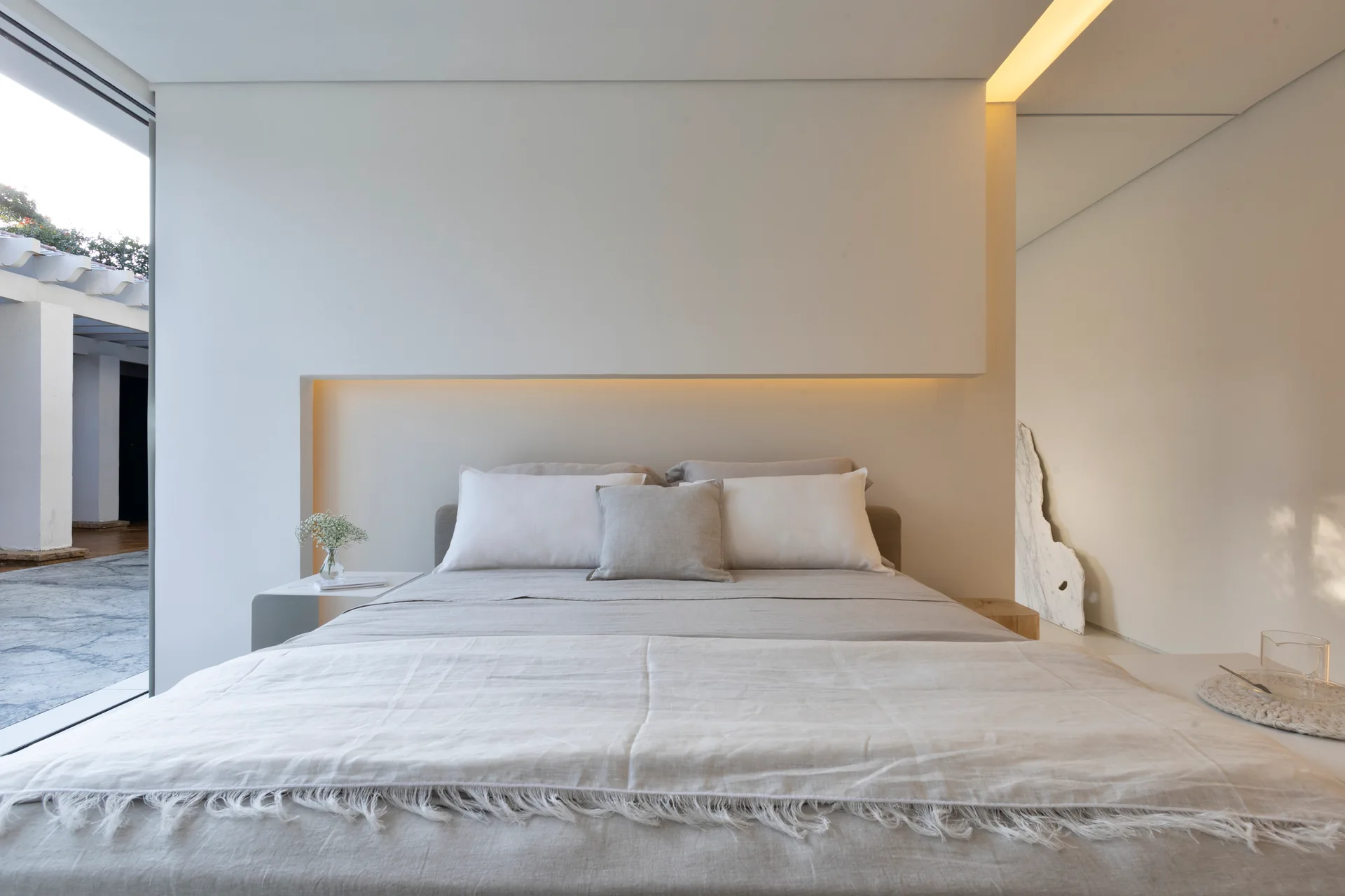 A minimalist bedroom featuring neutral linen bedding against a white wall with an integrated lighted niche and a large side window.