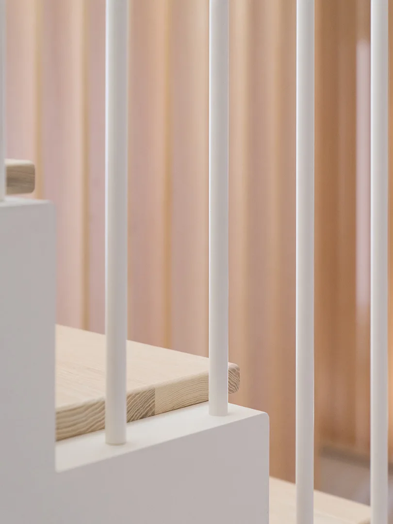 A close-up view of a minimalist staircase featuring slender white vertical balusters, light wood treads, and a warm wood-paneled background.