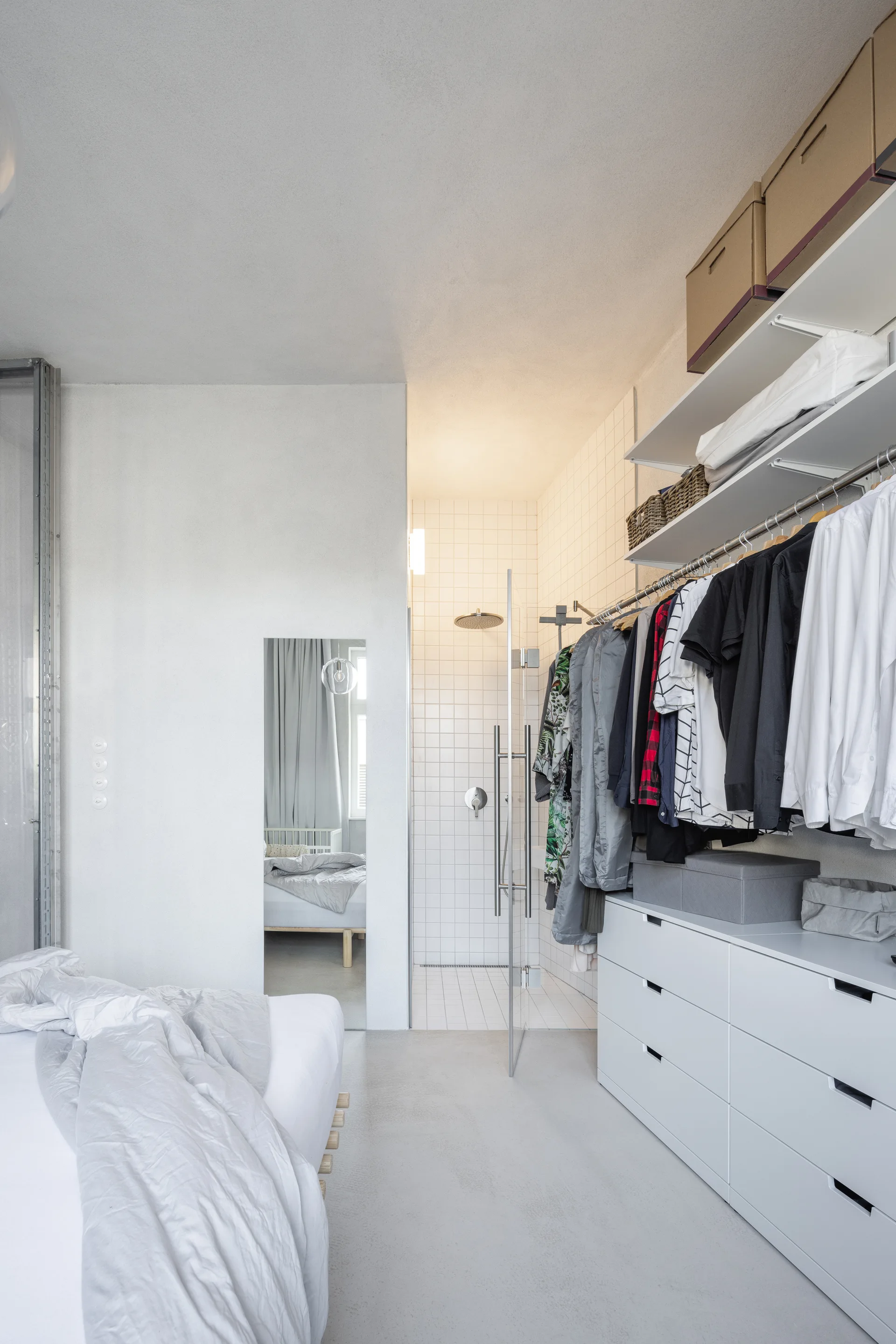 A minimalist bedroom featuring an open closet wardrobe next to a glass-enclosed shower with white grid tiles.