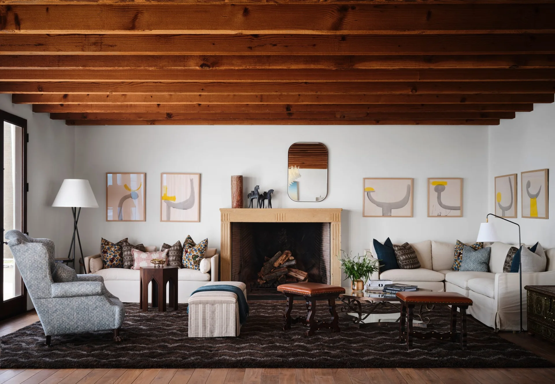 A living room features exposed wood ceiling beams, a stone fireplace, cream sofas, a patterned rug, and abstract art framed on the walls.