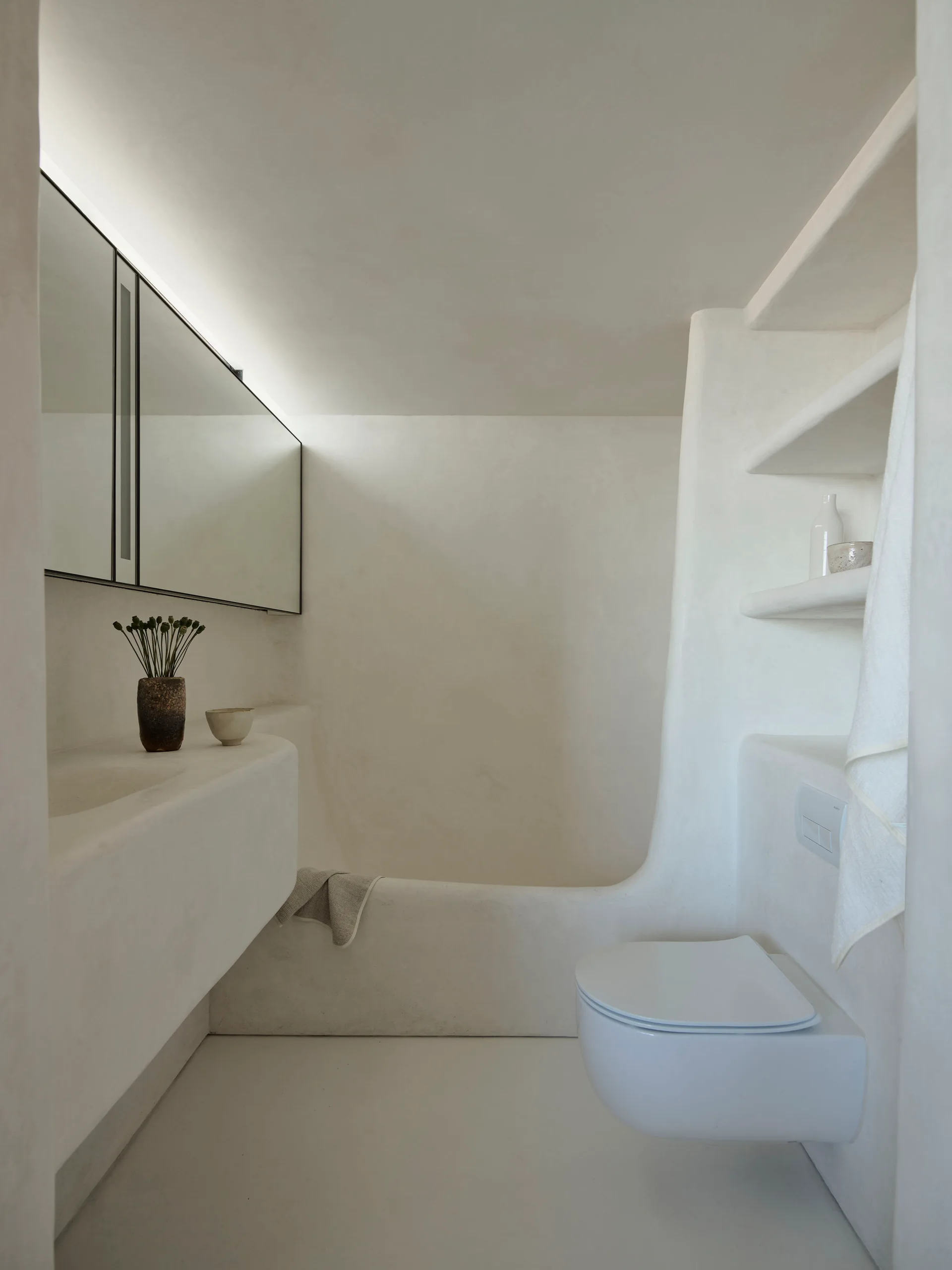 A minimalist bathroom features seamless white plaster surfaces, an integrated curved vanity basin, built-in shelving, and a wall-hung toilet.