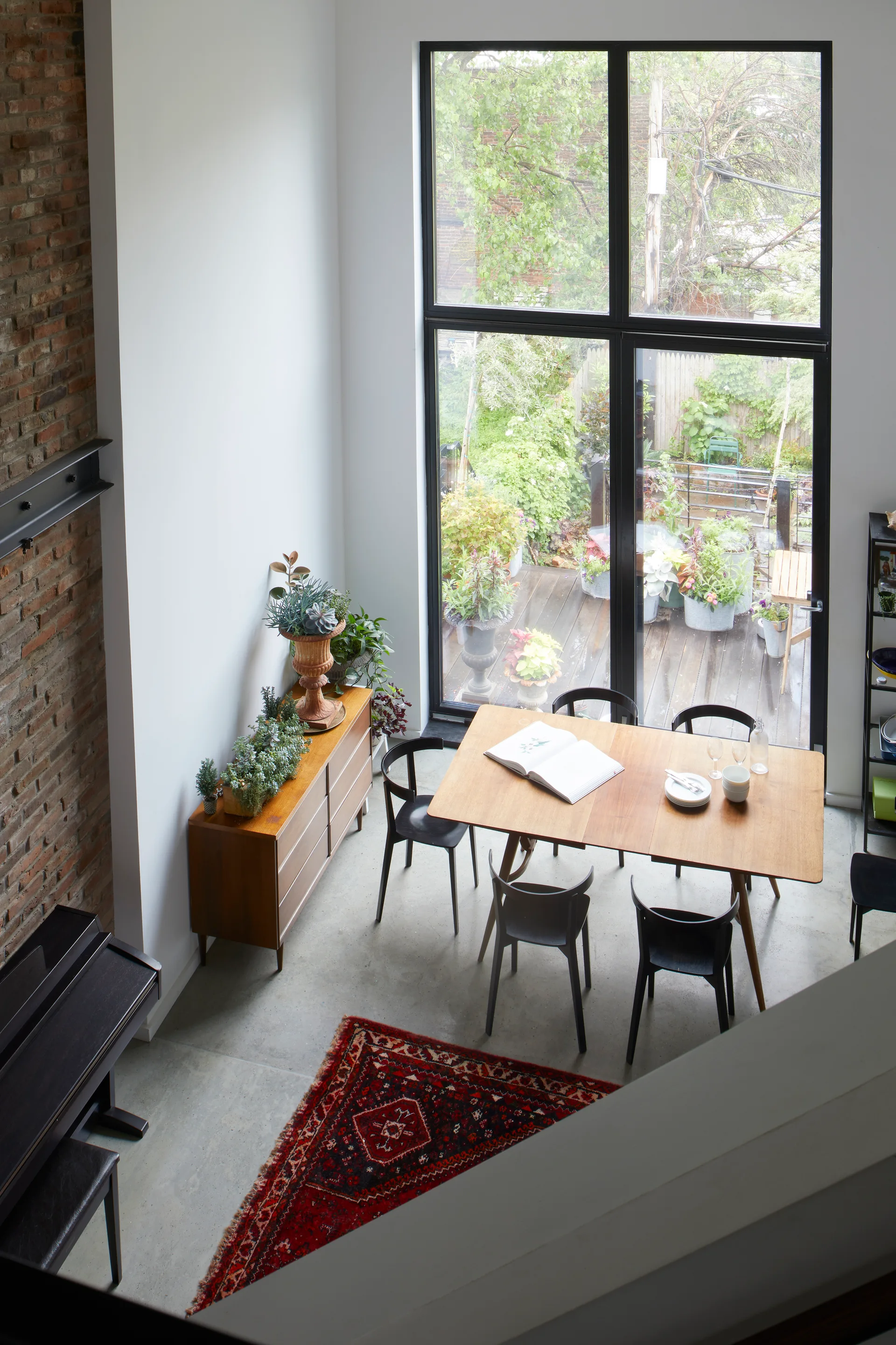 A high-angle view of a dining room with a wooden table, black chairs, a vintage red rug on concrete flooring, and a large black-framed window.