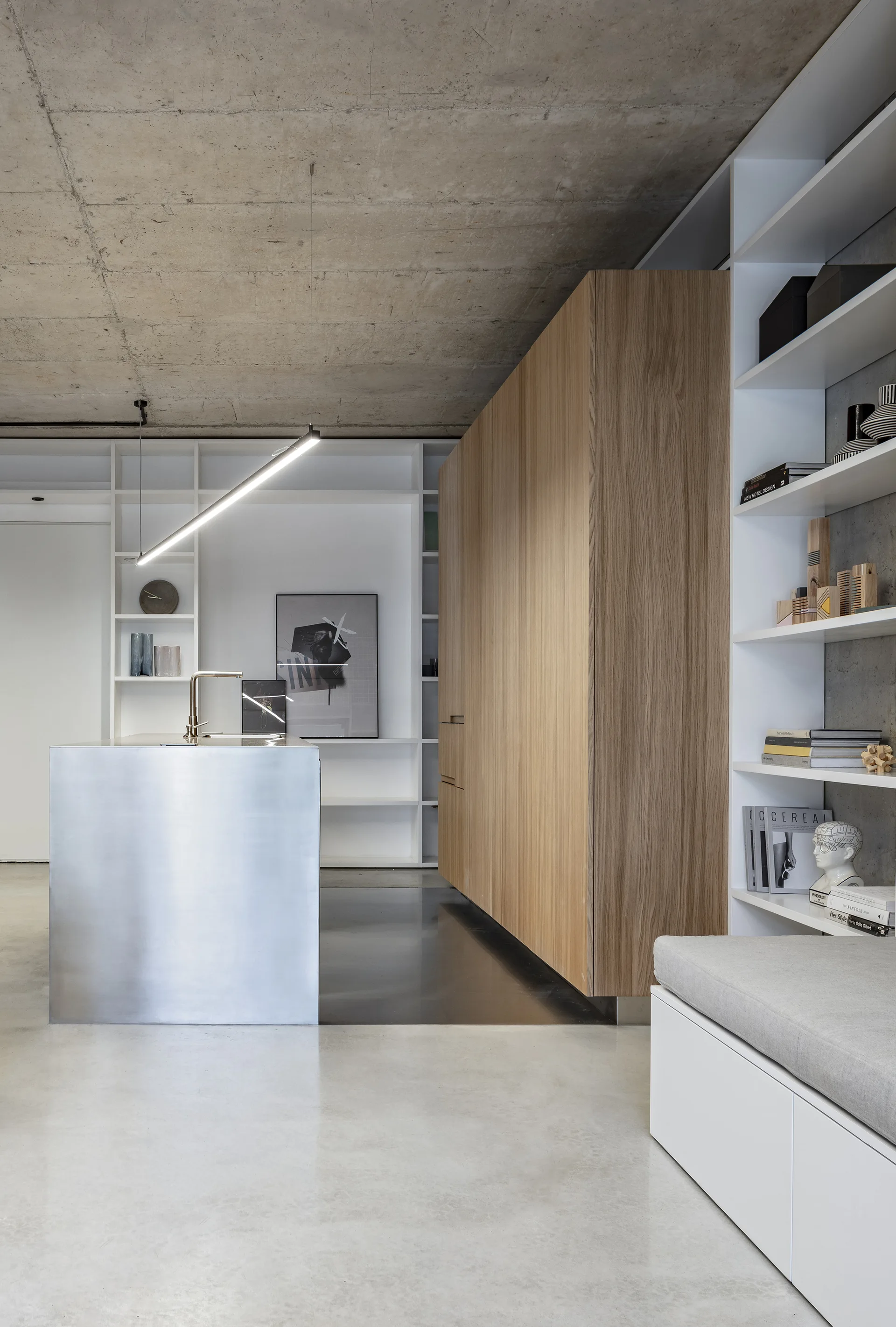 A modern kitchen features a stainless steel island, tall wood-grain cabinets, built-in white shelving, and an exposed concrete ceiling.