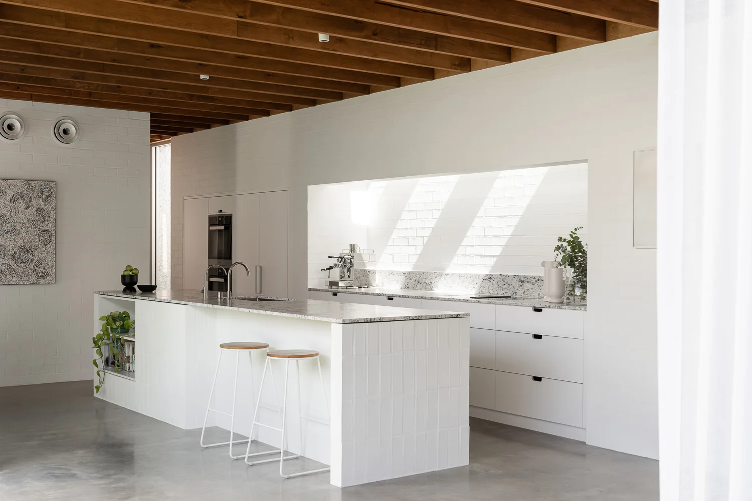A minimalist white kitchen features a tiled island with bar stools, speckled stone countertops, and exposed wood ceiling beams over a concrete floor.