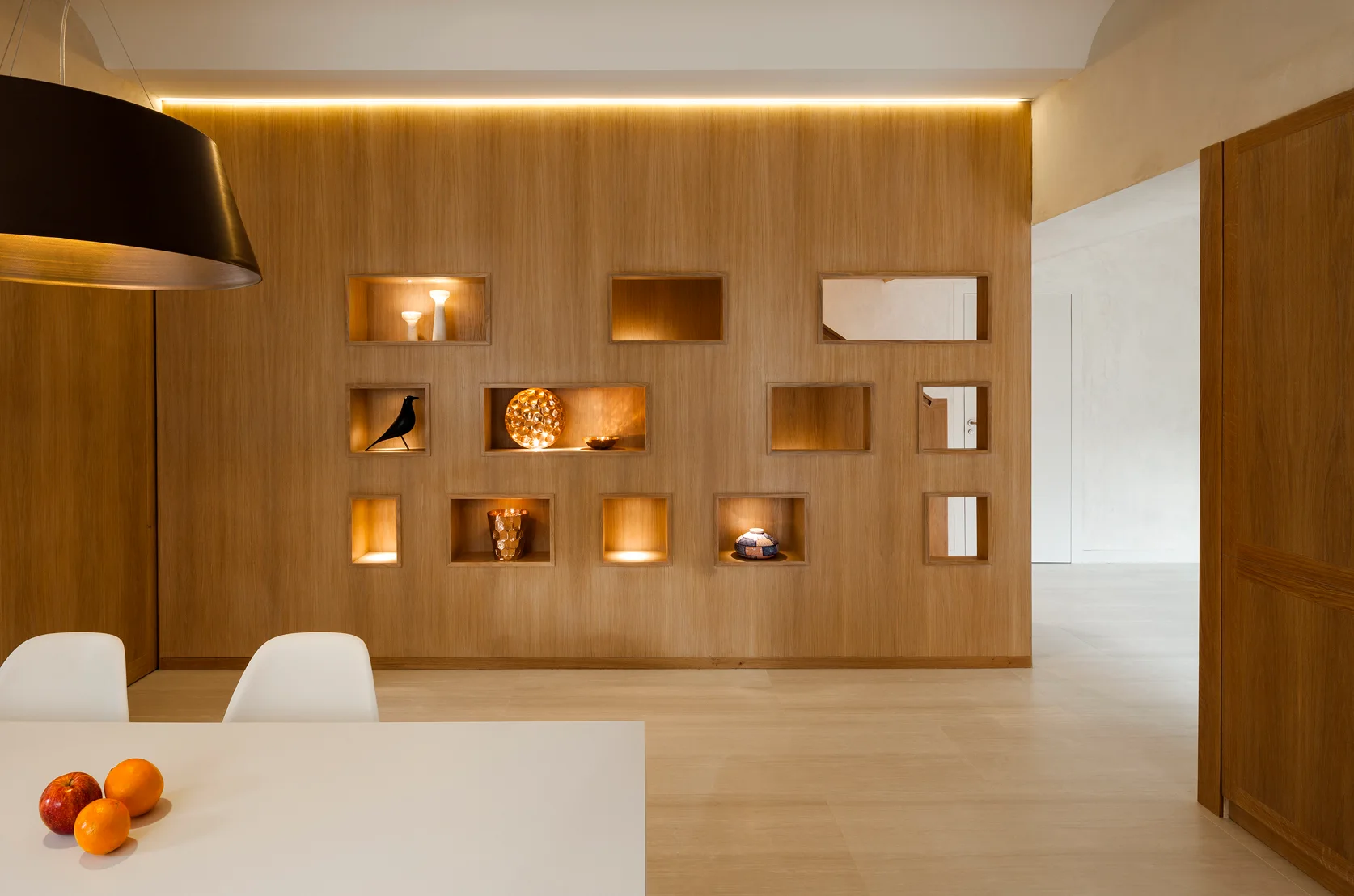 A modern dining room featuring a large wood-paneled wall with lit recessed display niches, a white table with fruit, and a black pendant light.