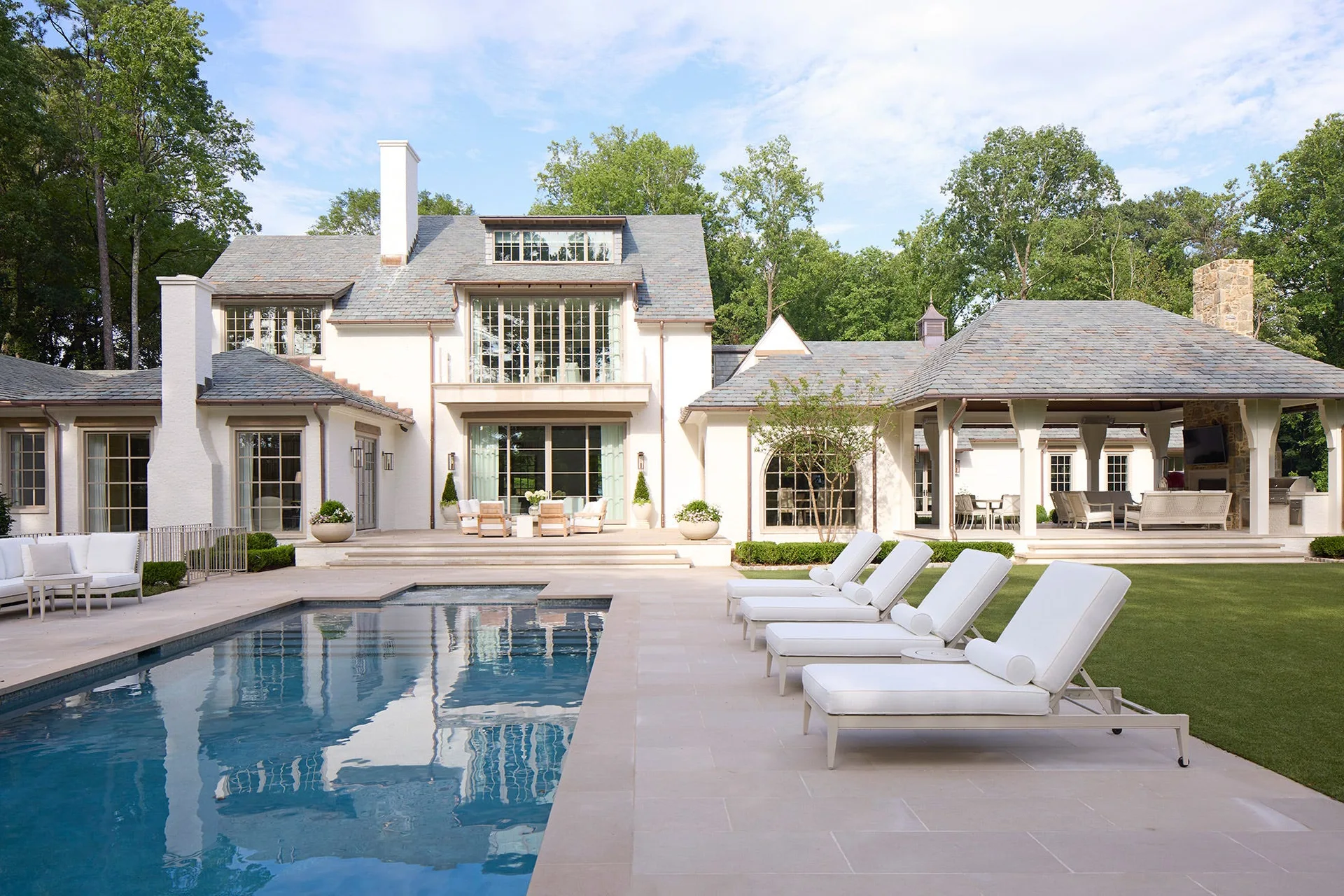 Four white lounge chairs sit on a light stone patio beside a large backyard swimming pool behind a white traditional home.