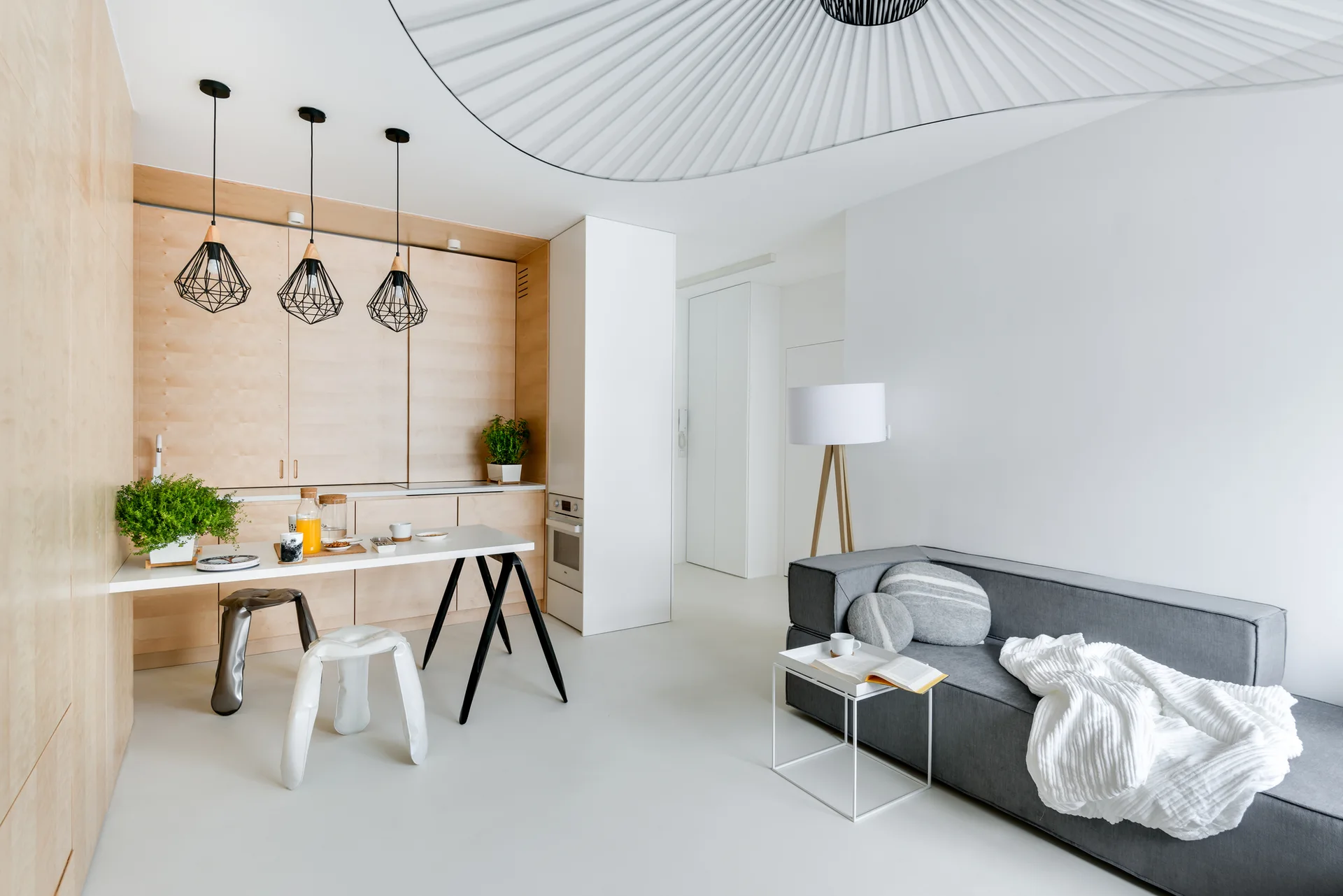 A minimalist open-concept living area with a grey sofa, integrated light wood kitchen cabinetry, and black geometric pendant lights.