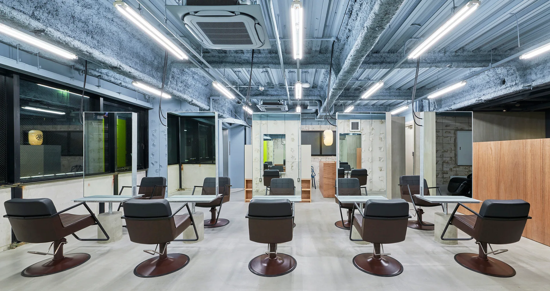 An industrial hair salon features minimalist styling stations with brown leather chairs, tall rectangular mirrors, and an exposed light blue ceiling.