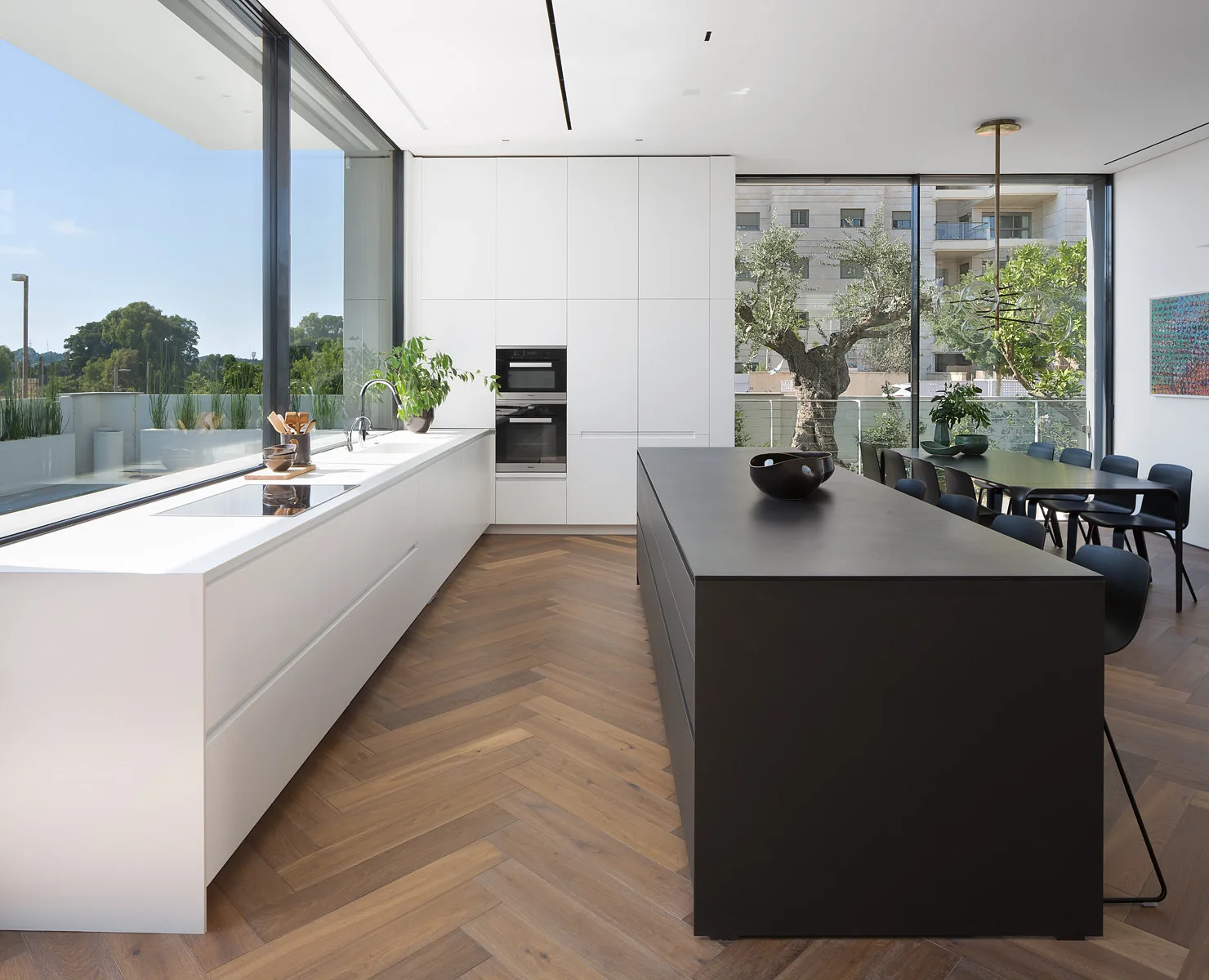 A modern kitchen featuring a large matte black island, white flat-panel cabinetry, floor-to-ceiling windows, and warm wood herringbone flooring.
