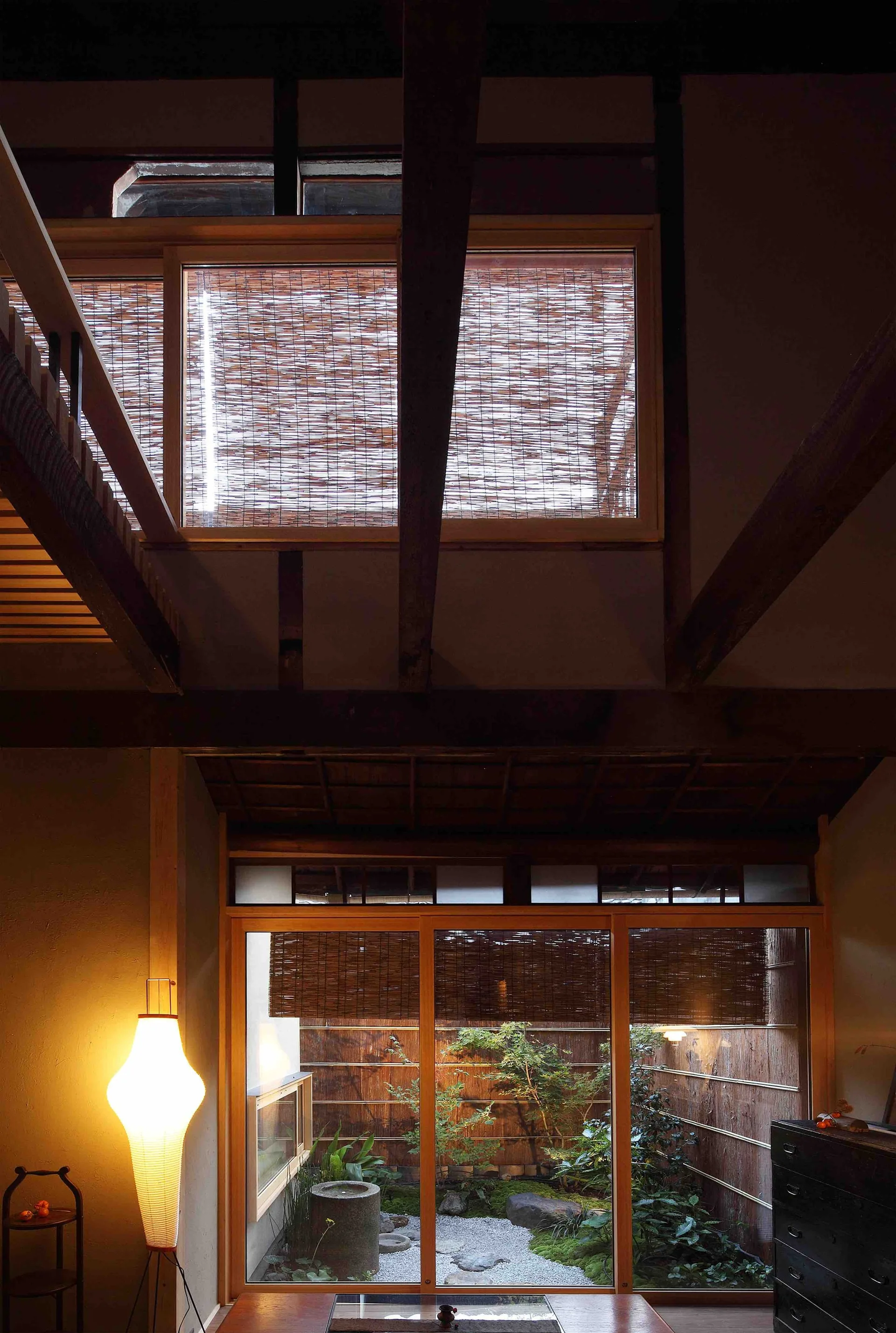 A Japanese living room featuring dark exposed timber ceiling beams, a glowing paper floor lamp, and glass doors looking out onto a small courtyard garden.