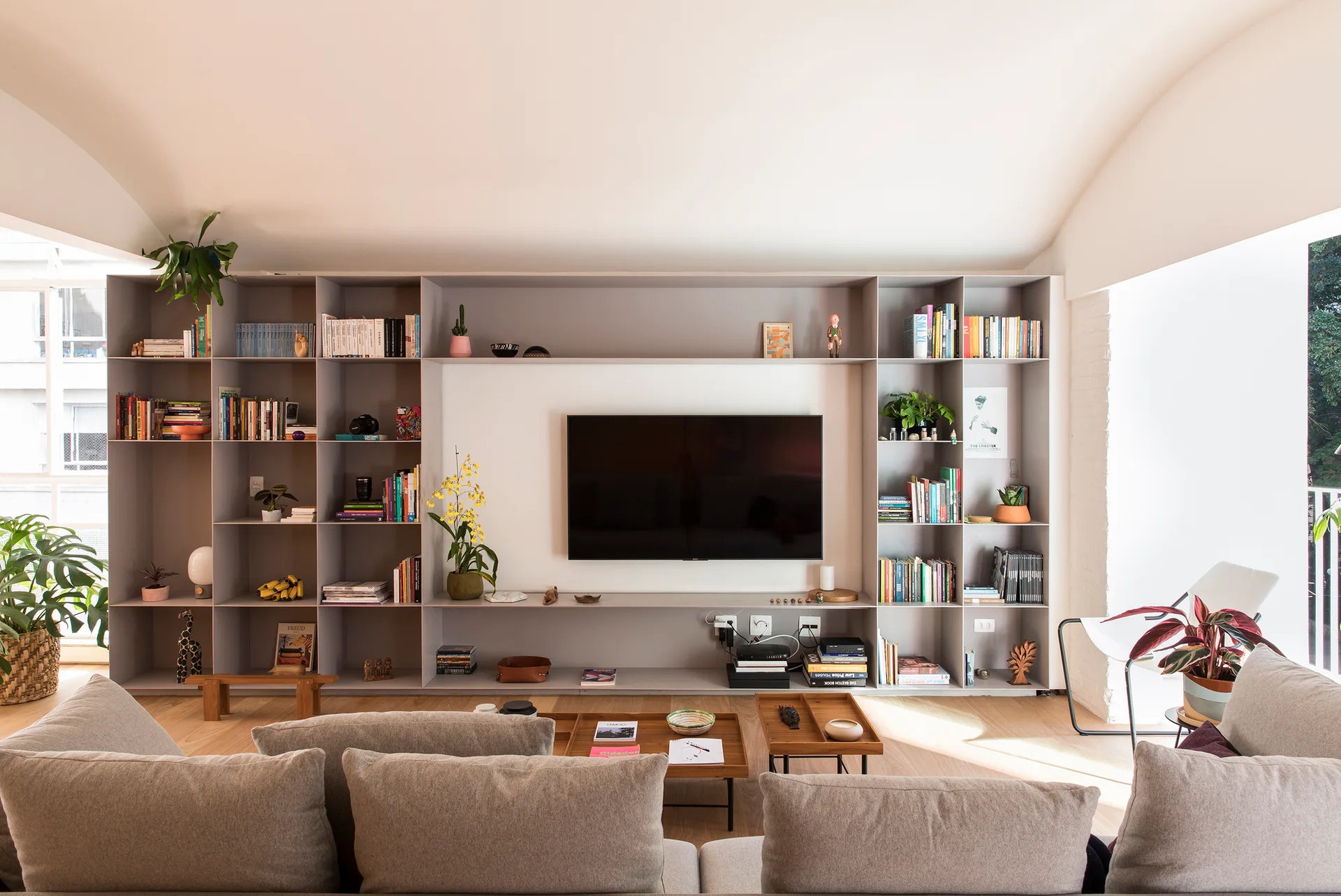 A bright living room features grey upholstered sofas facing a large, wall-to-wall light grey built-in shelving unit with an integrated TV and books.