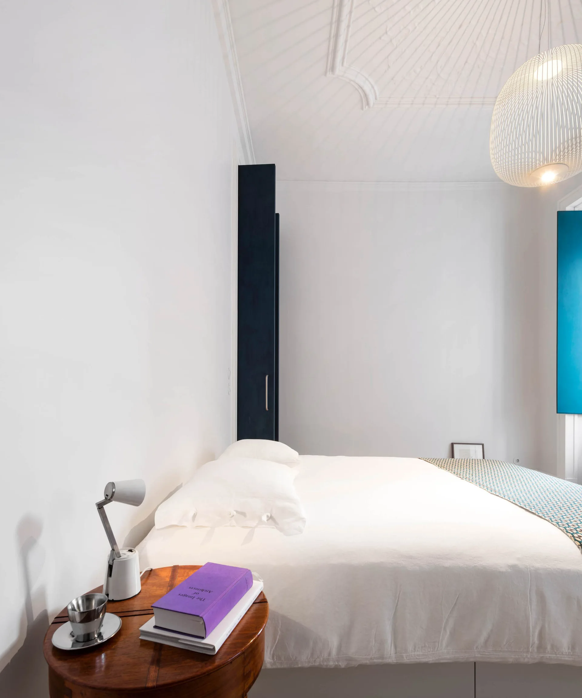 A bright white bedroom with a white-dressed bed, an ornate plaster ceiling, a round wooden nightstand, and a tall dark blue wardrobe.