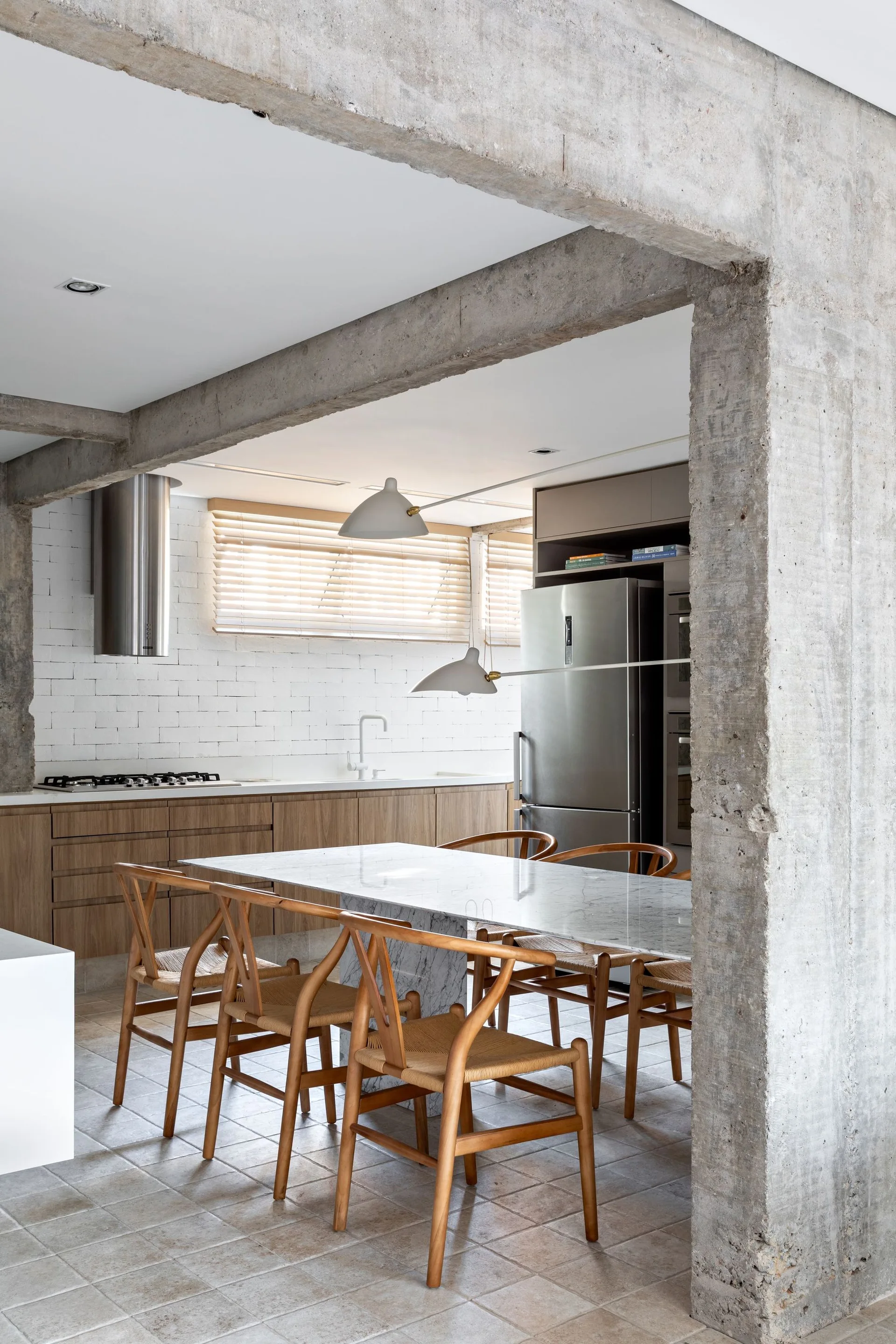 A modern dining area with a marble-topped table and wood wishbone chairs set against exposed concrete columns and white brick walls.