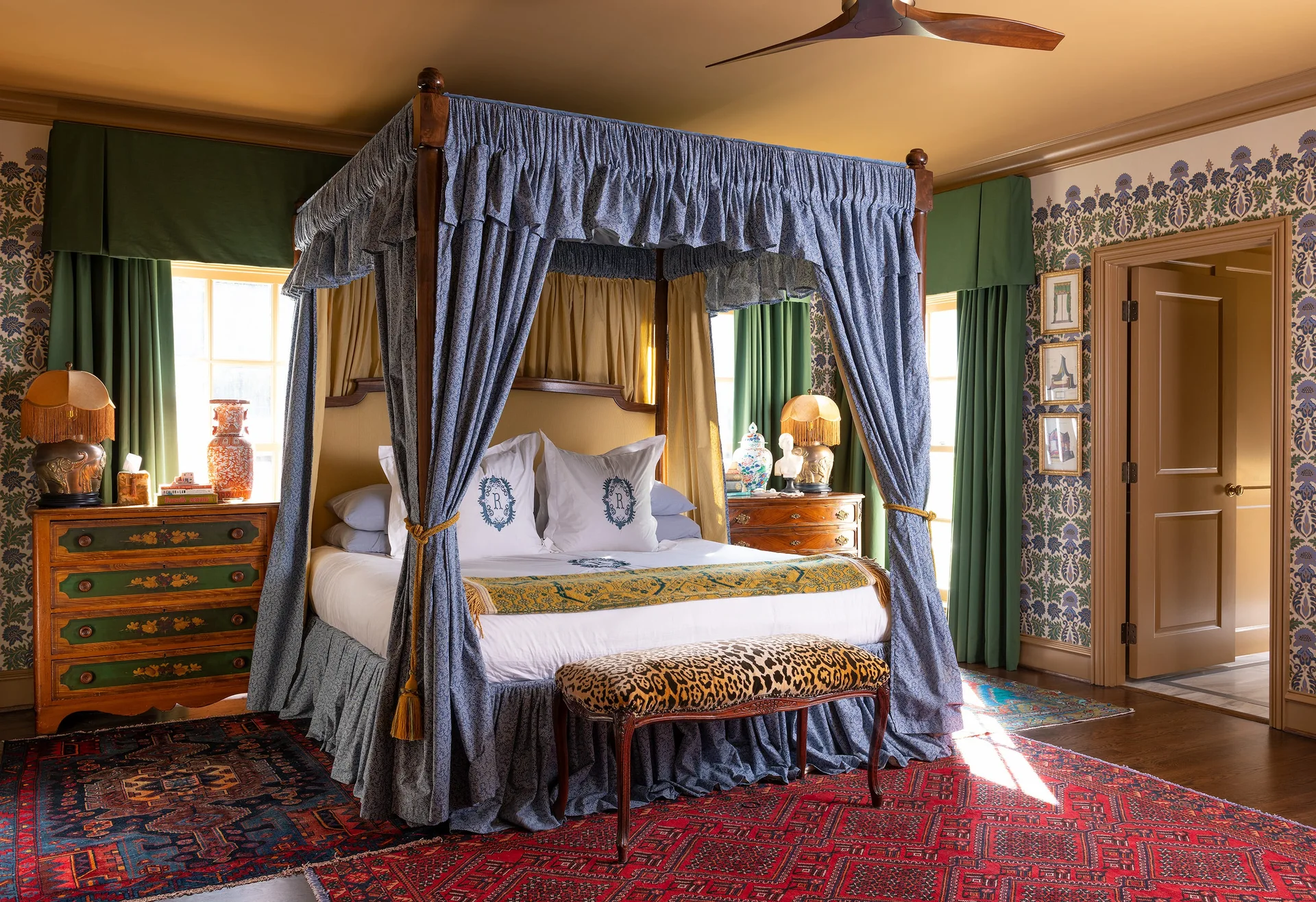 A traditional bedroom featuring a blue patterned canopy bed, intricate botanical wallpaper, a leopard-print bench, and a red patterned area rug.