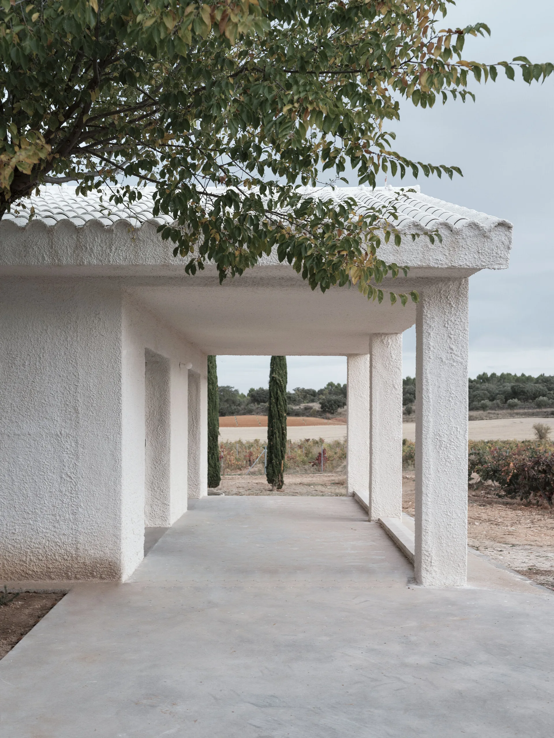 A covered concrete porch features white textured stucco columns and matching roof tiles framing a view of a rural landscape.