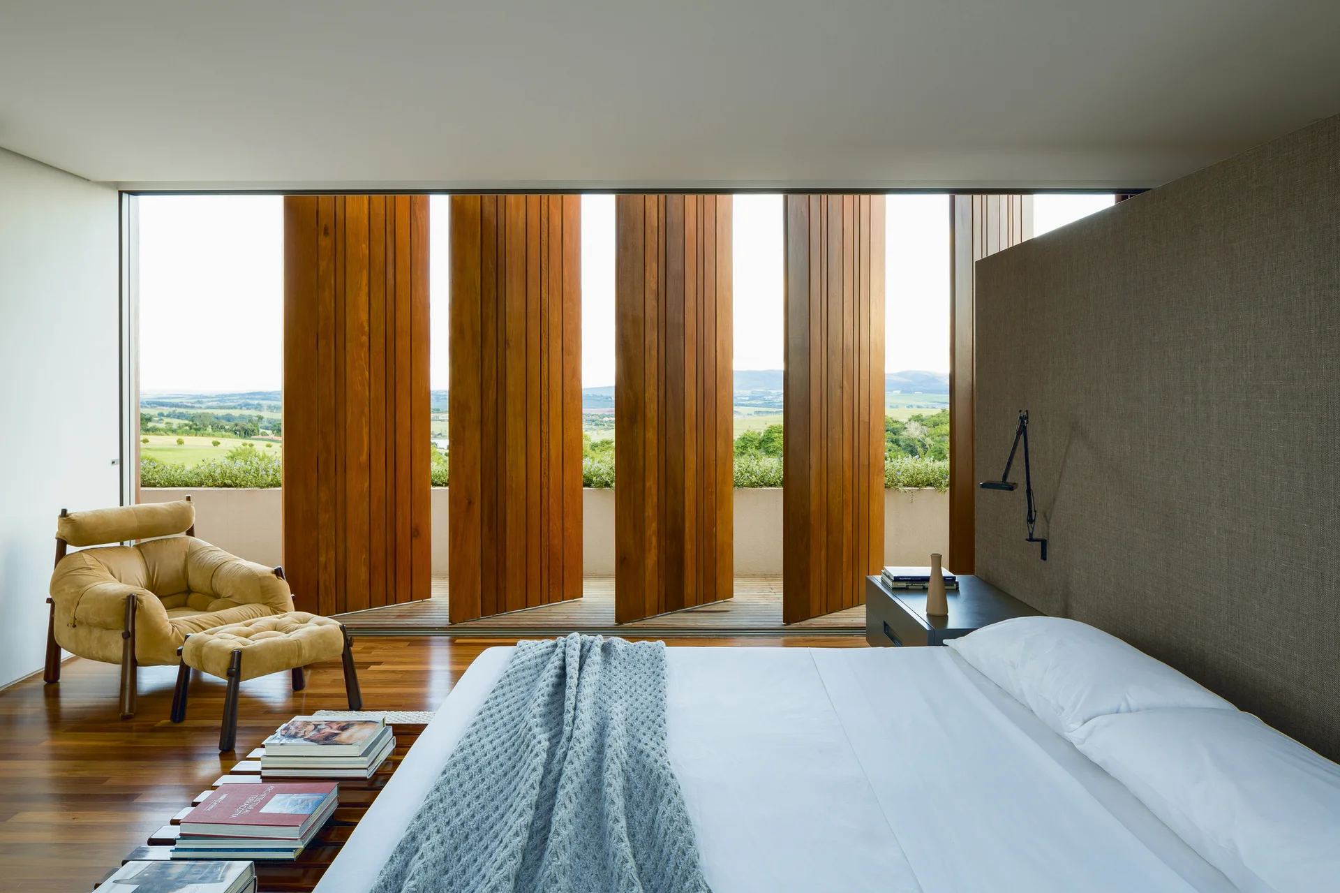 A modern bedroom features a bed with white linens, a tan lounge chair, and large vertical wood shutters opening to an outdoor terrace.
