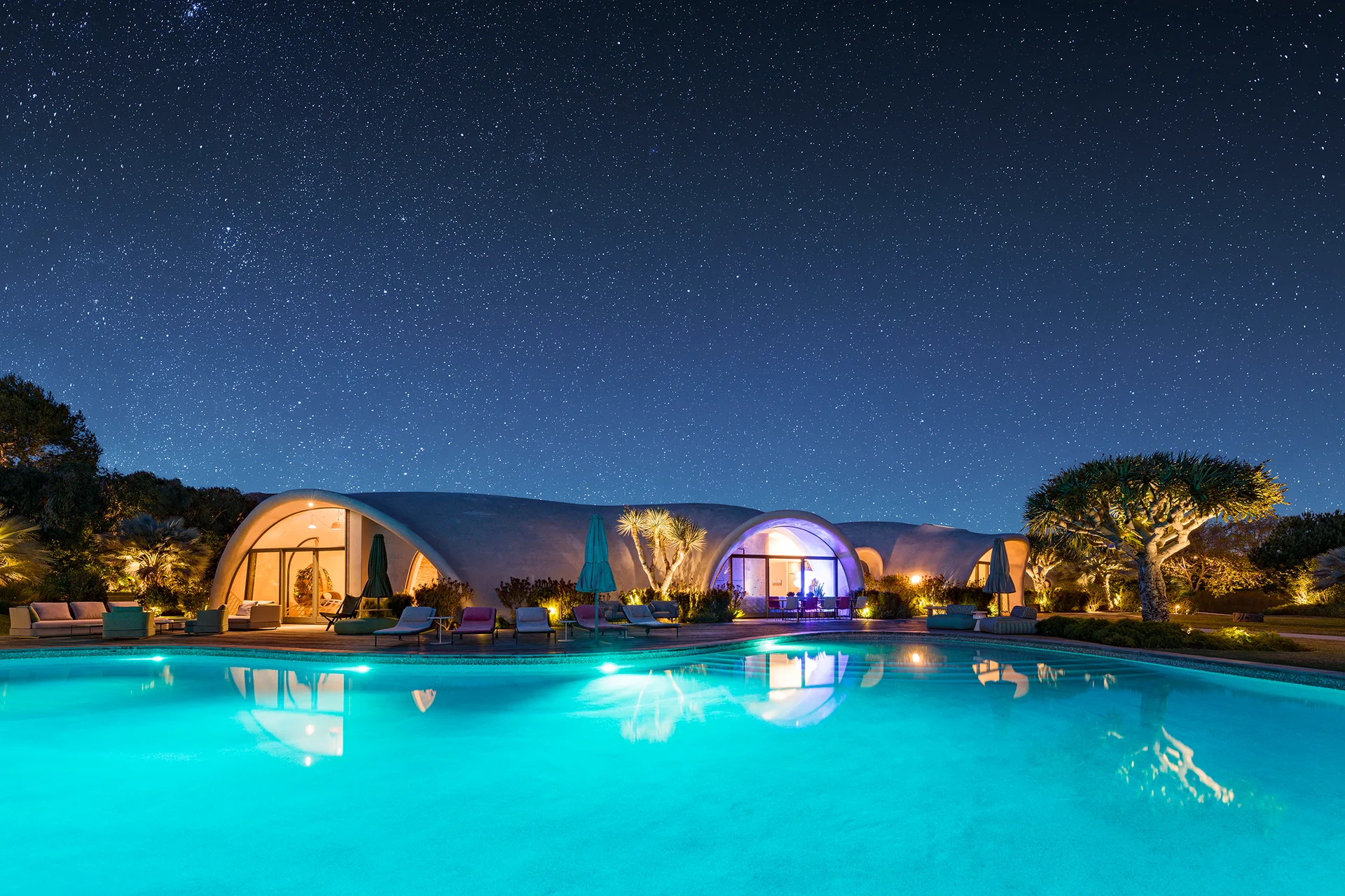 Vaulted concrete dome structures sit beside a large glowing turquoise swimming pool on a wooden deck under a starry night sky.