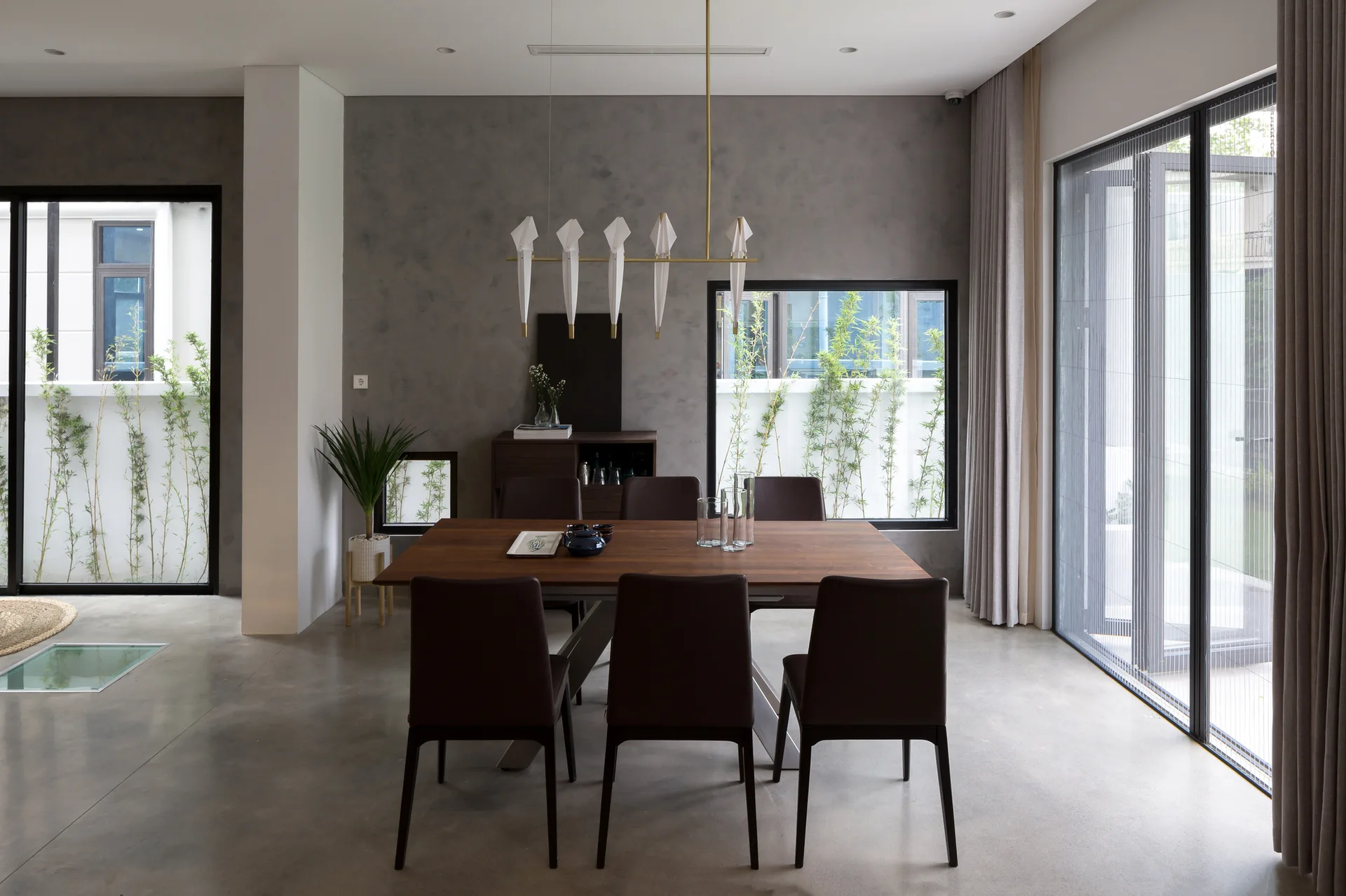 A modern dining room features a dark wood table, brown leather chairs, a bird-shaped pendant light, and a textured grey plaster wall with windows.