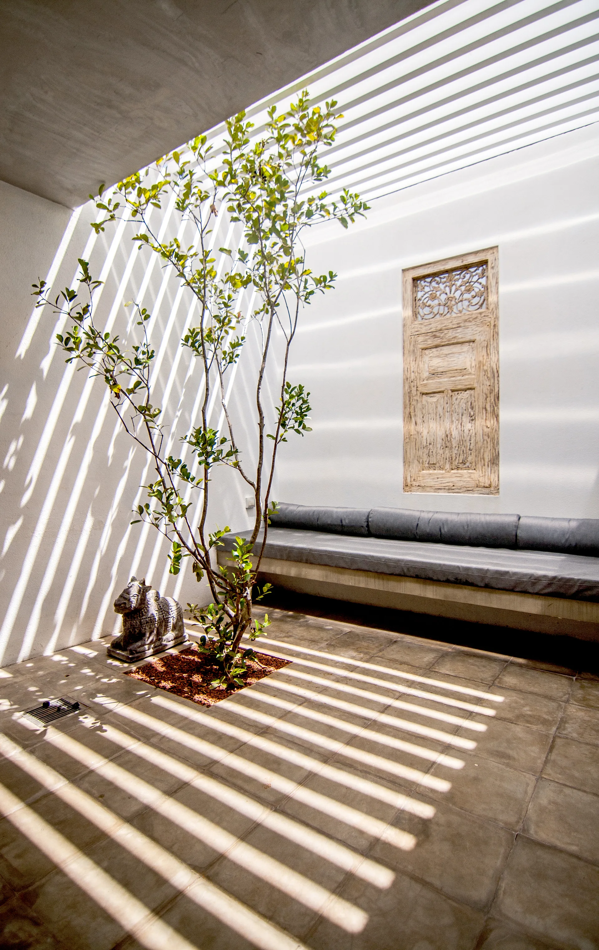 A minimalist outdoor courtyard features a built-in bench with grey cushions, a small tree, and strong linear shadows cast by a slatted roof overhead.