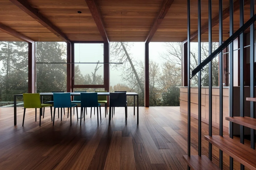 A modern dining room features a wood-paneled floor and ceiling, a dark table with teal and green chairs, and large windows looking out on trees.