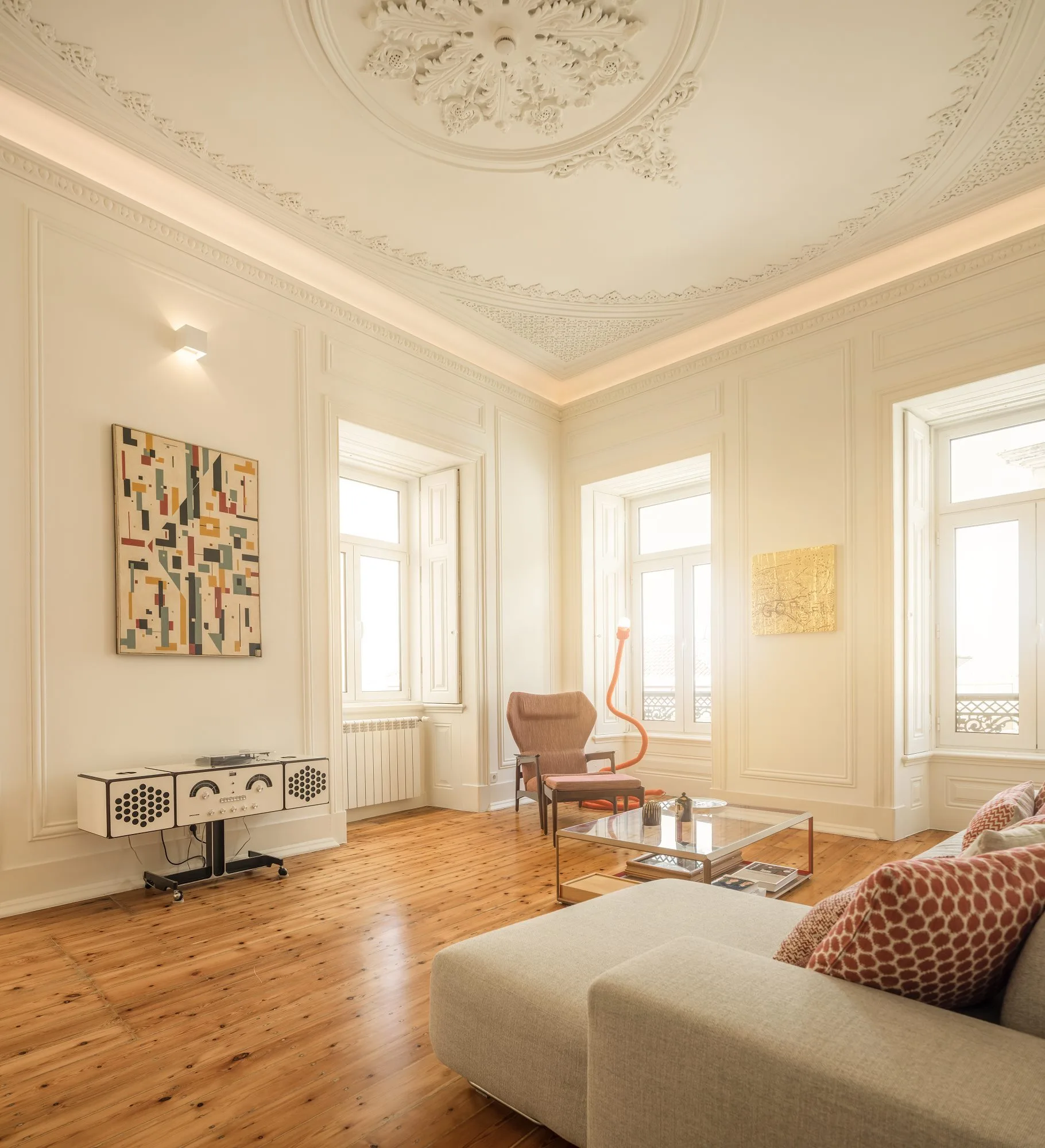 A bright living room features high ceilings with an ornate plaster medallion, tall windows, a light-colored sofa, and warm pine wood floors.