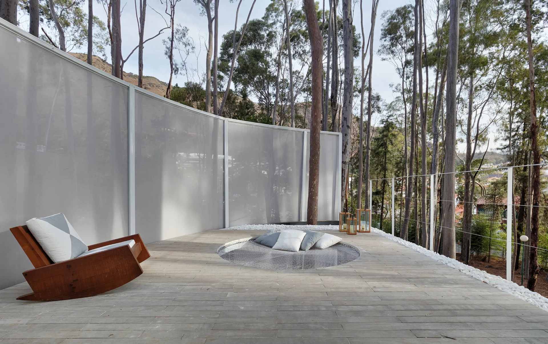 A wooden outdoor patio deck features a wooden rocking chair, a sunken circular seating net filled with cushions, and a curved white privacy screen.
