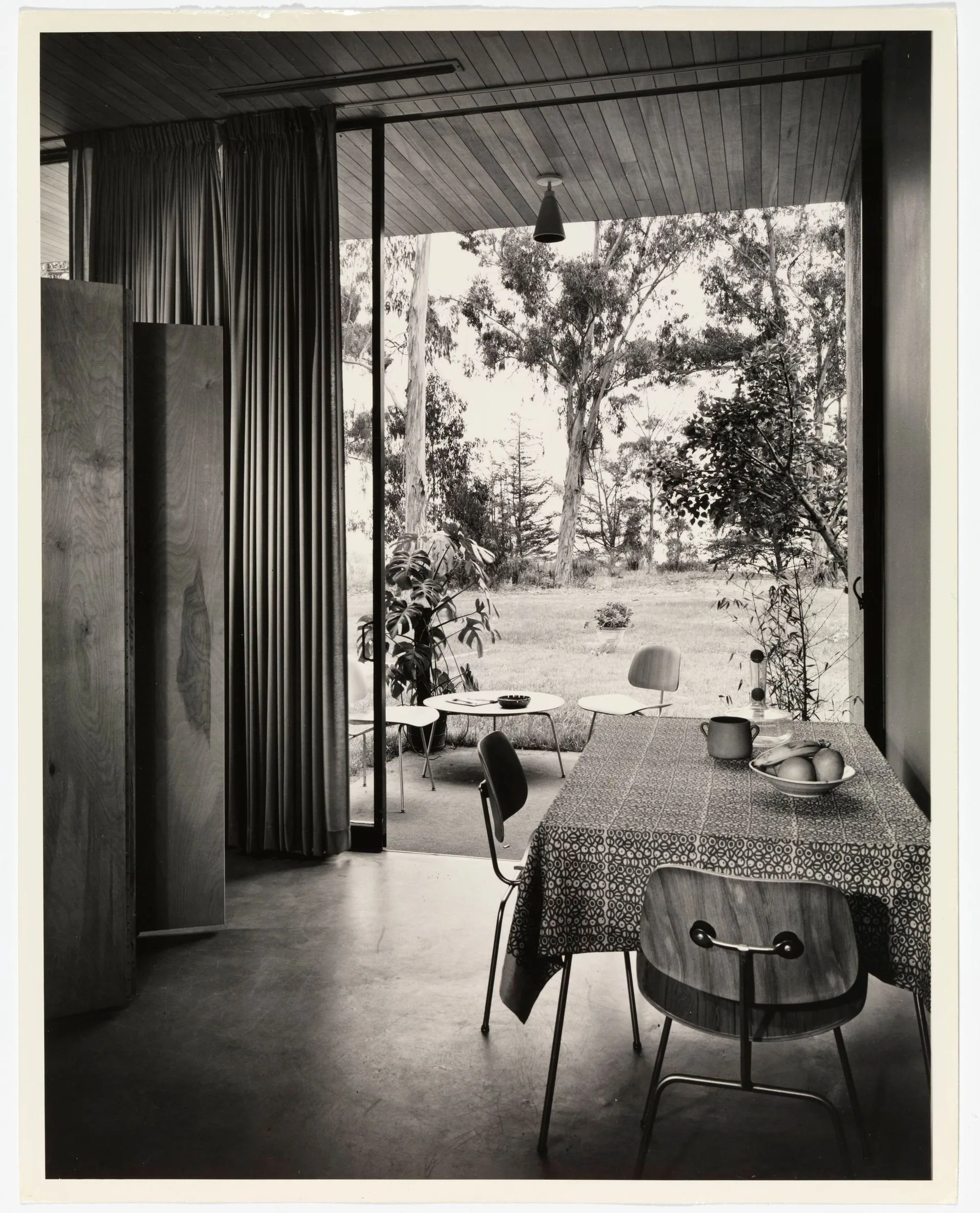 A black and white photo of a dining room with molded plywood chairs and a patterned tablecloth next to a large sliding glass door opening to a patio.