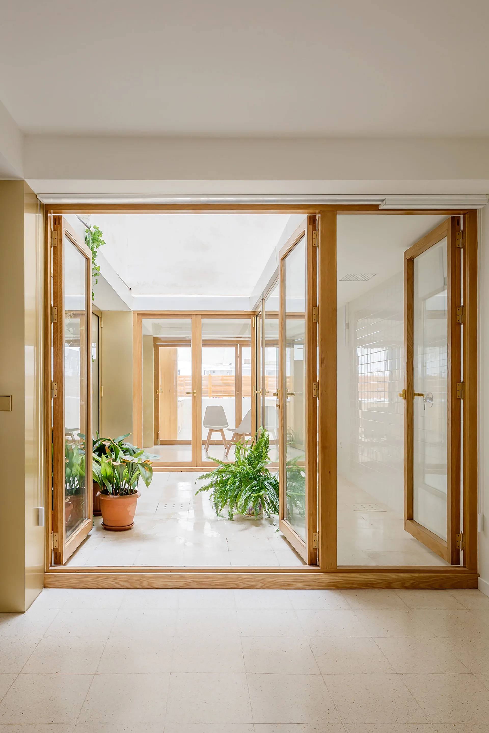 Wood-framed folding glass doors open into a bright indoor atrium featuring white tiled floors, a skylight, and several green potted plants.