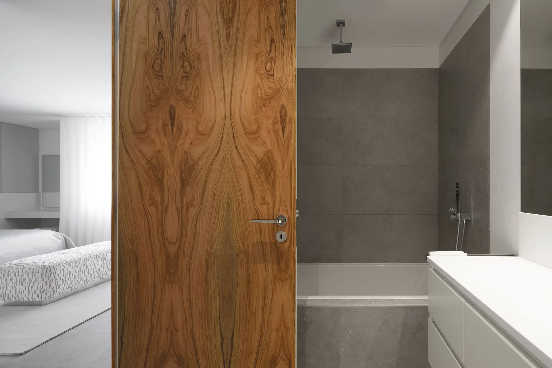A modern bathroom with grey tiled walls, a white vanity, and a large wood grain door looking into a minimalist bedroom.