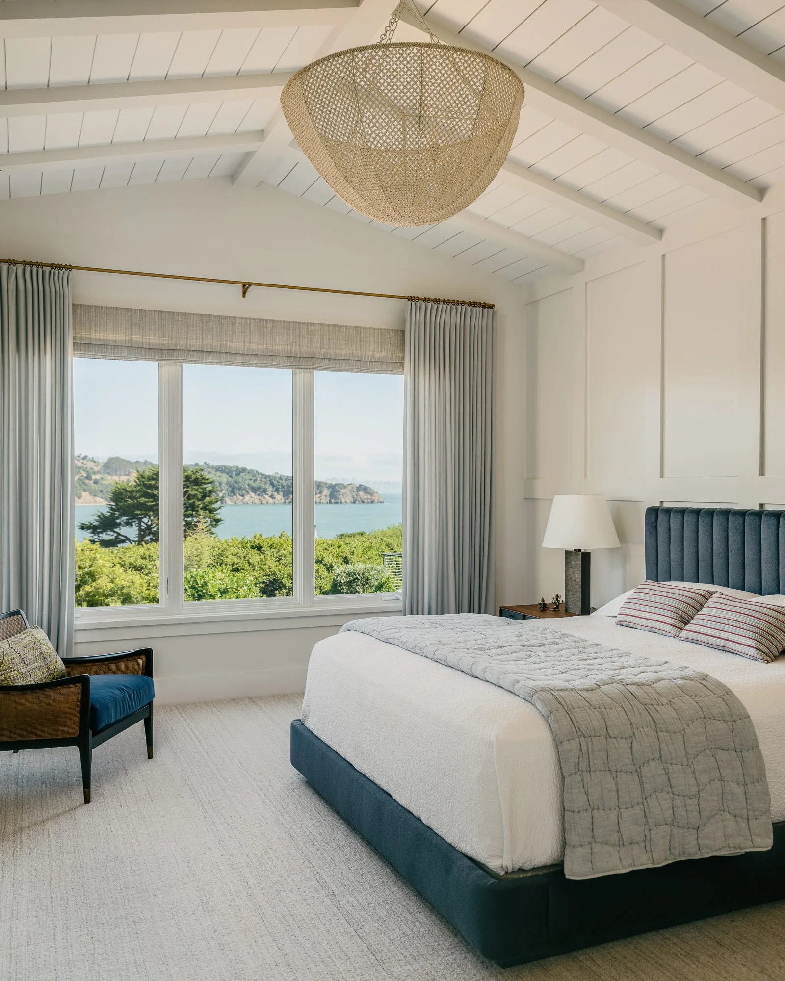 A light-filled bedroom featuring a navy upholstered bed, a large woven pendant light, and a window with scenic coastal views.