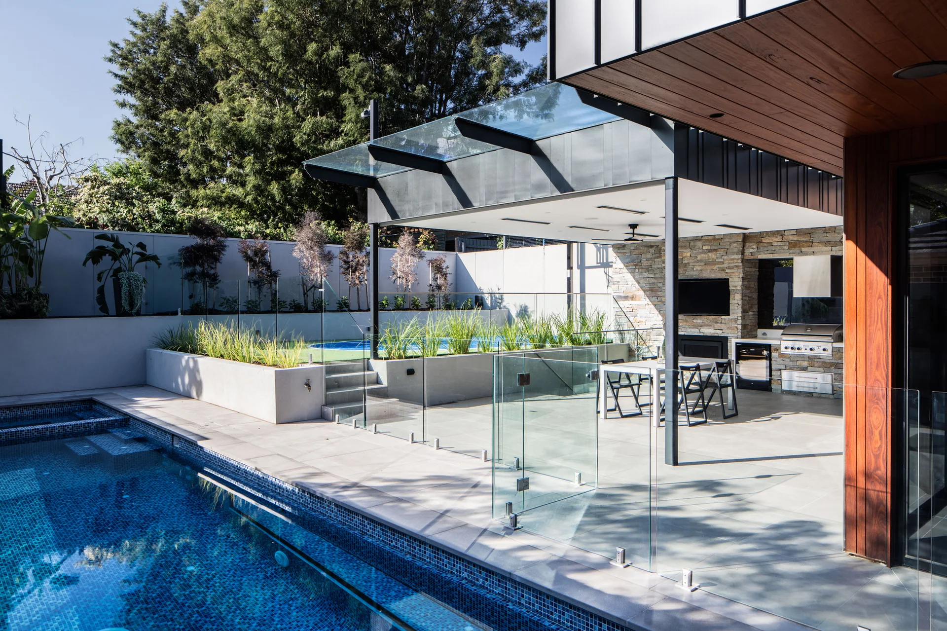 A modern backyard patio features a tiled swimming pool enclosed by a frameless glass fence next to a covered outdoor kitchen and dining area.