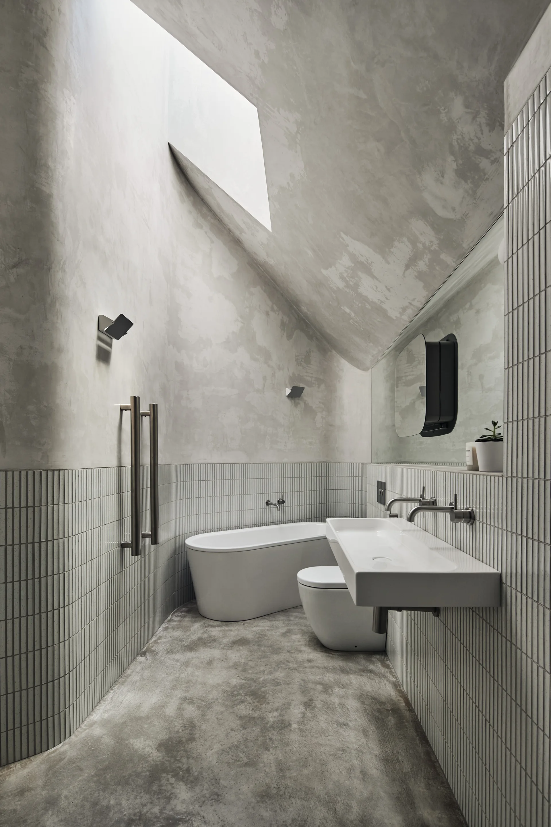A contemporary bathroom features a sloped ceiling with a skylight, vertical white tile walls, a freestanding tub, and concrete flooring.