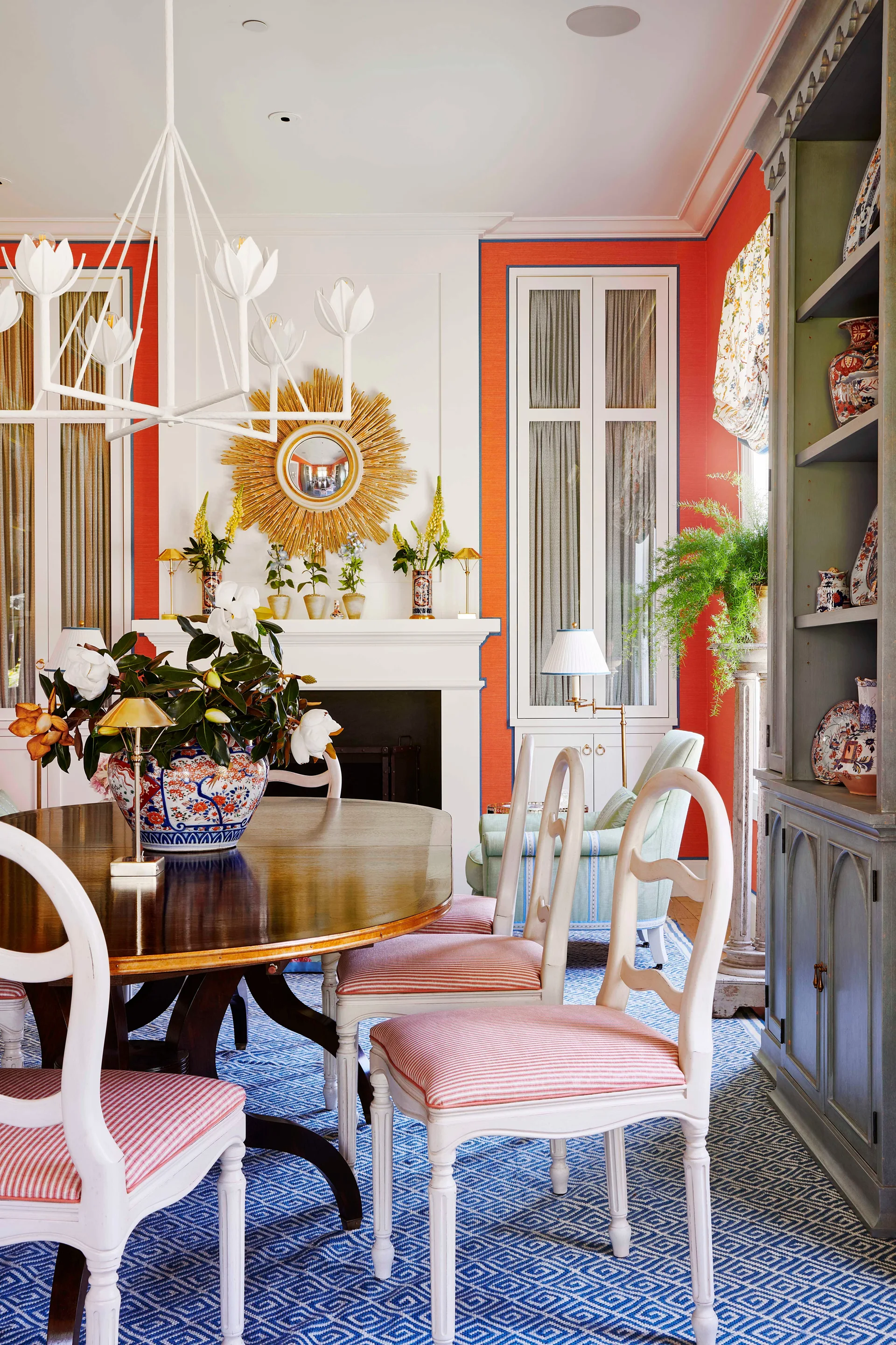 A traditional dining room features a dark wood table with striped chairs, a blue geometric rug, and bright orange wall panels surrounding a white fireplace.