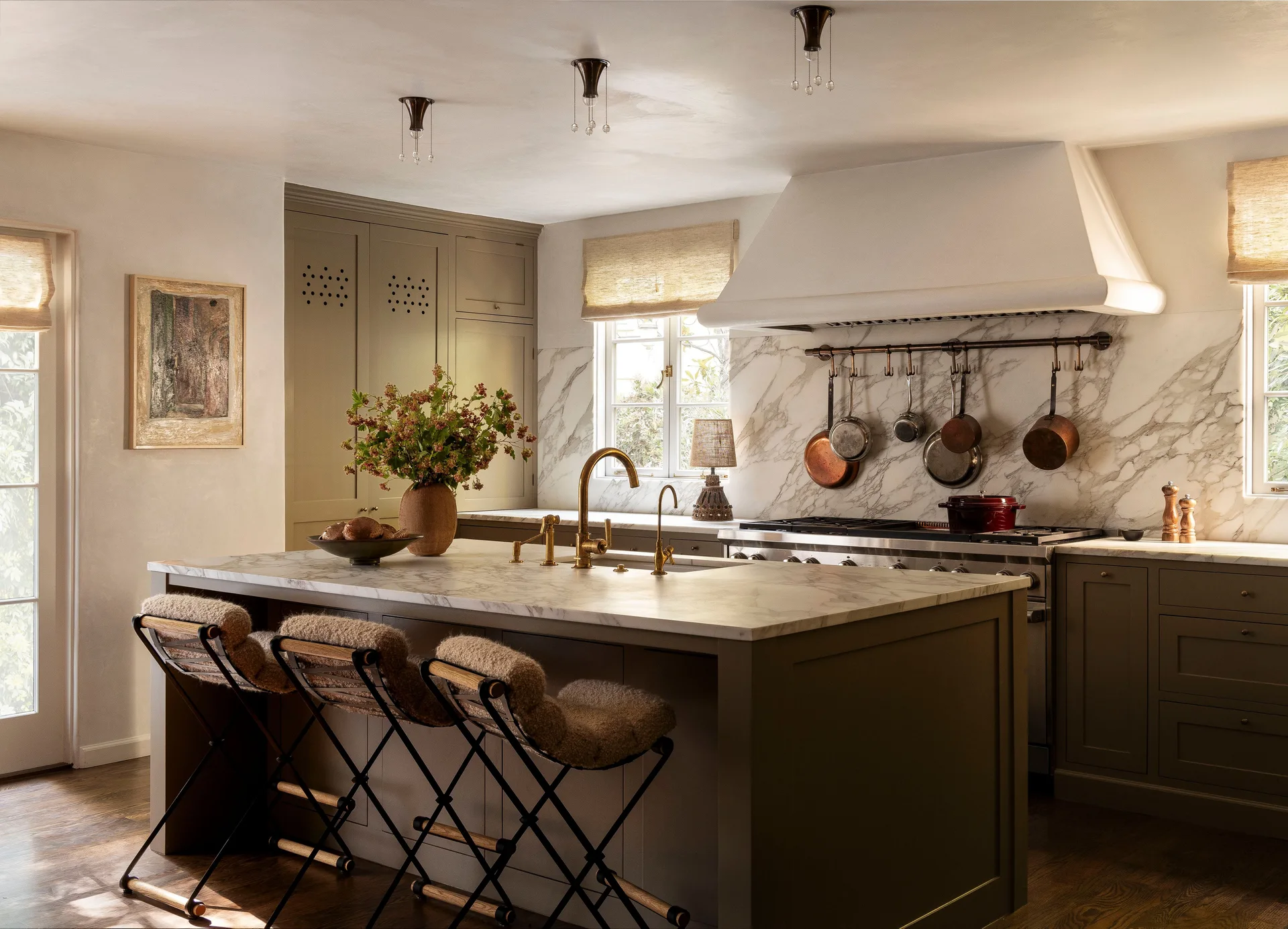 An olive green kitchen featuring a central island with a marble countertop, shearling-cushioned bar stools, and a matching marble slab backsplash.
