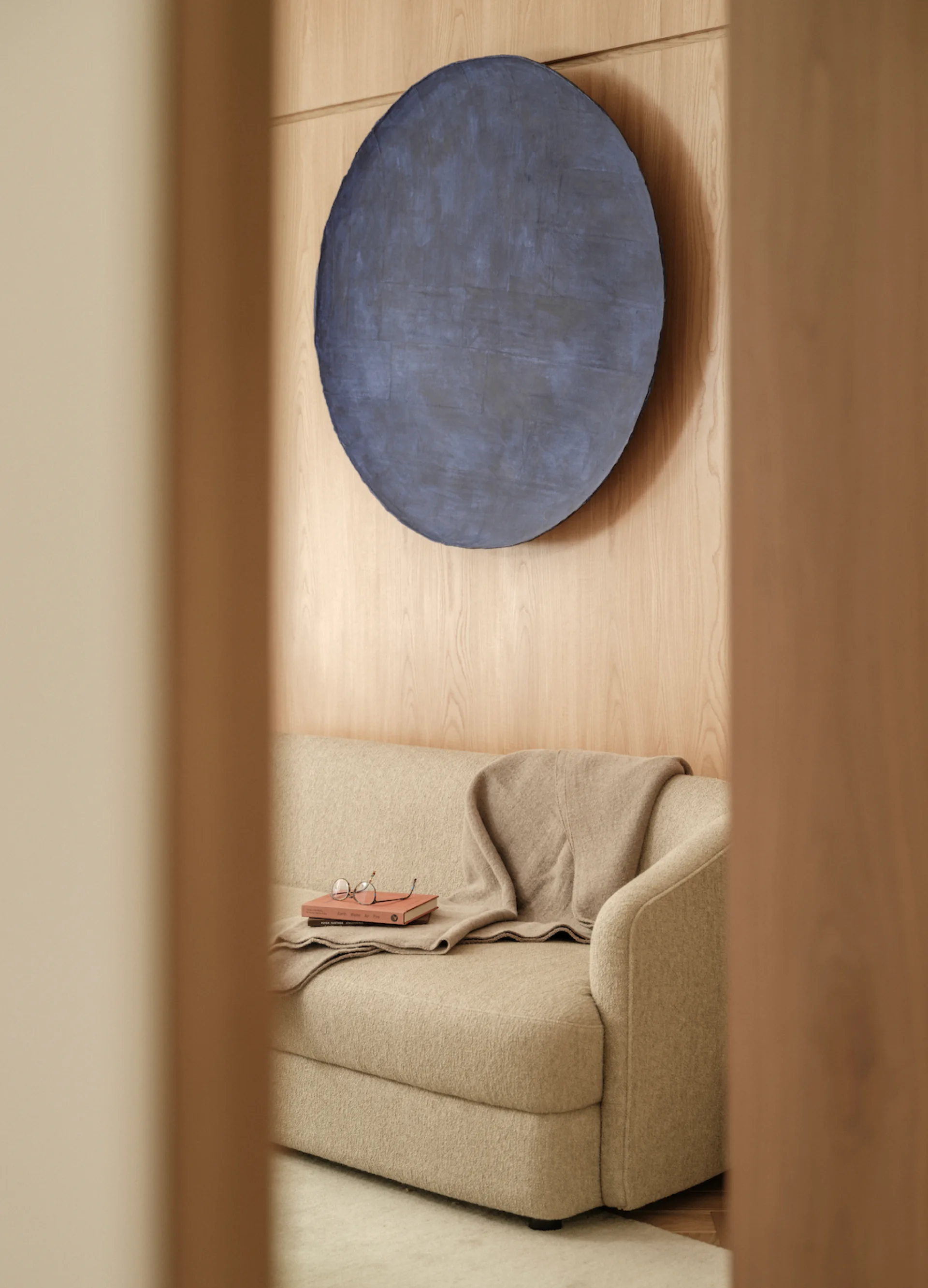 A beige sofa sits against a light wood paneled wall beneath a large circular blue painting, framed through a wooden doorway.