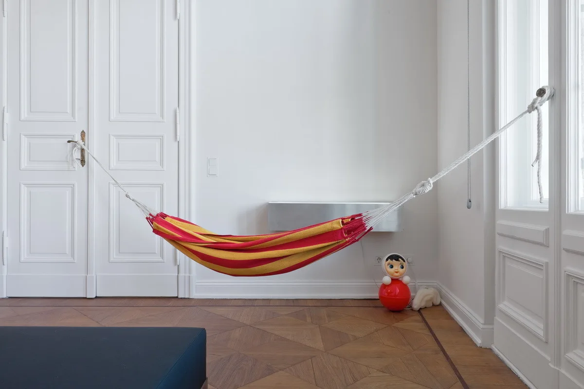 A red and yellow striped fabric hammock is suspended between a white paneled door and window frame in a room with parquet flooring.