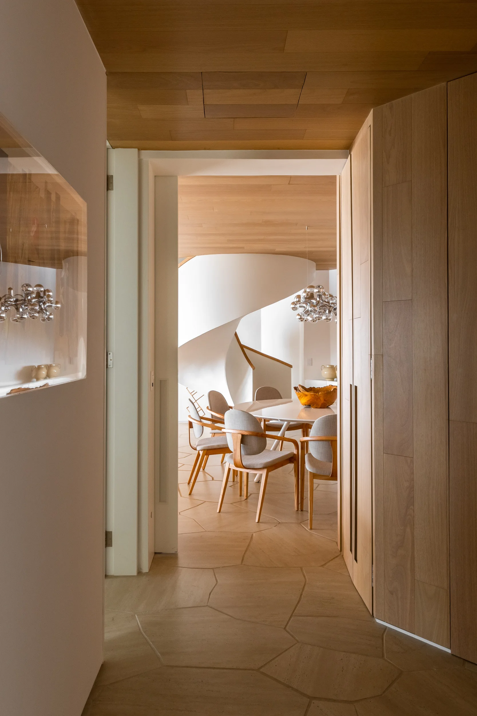 A view through a wood-paneled hallway looking into a dining room with a round table, upholstered chairs, and a white spiral staircase.